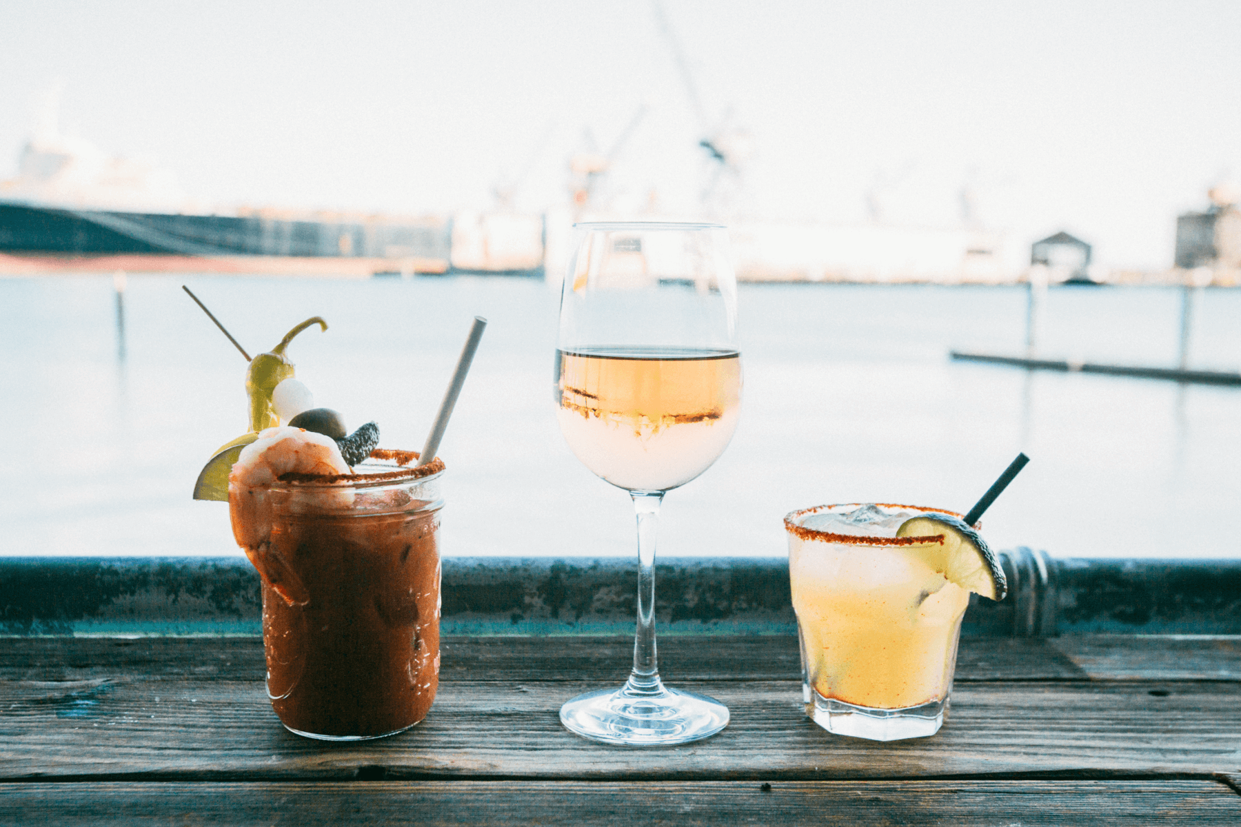 A bloody mary, a glass of wine, and a margarita on a bar overlooking water.
