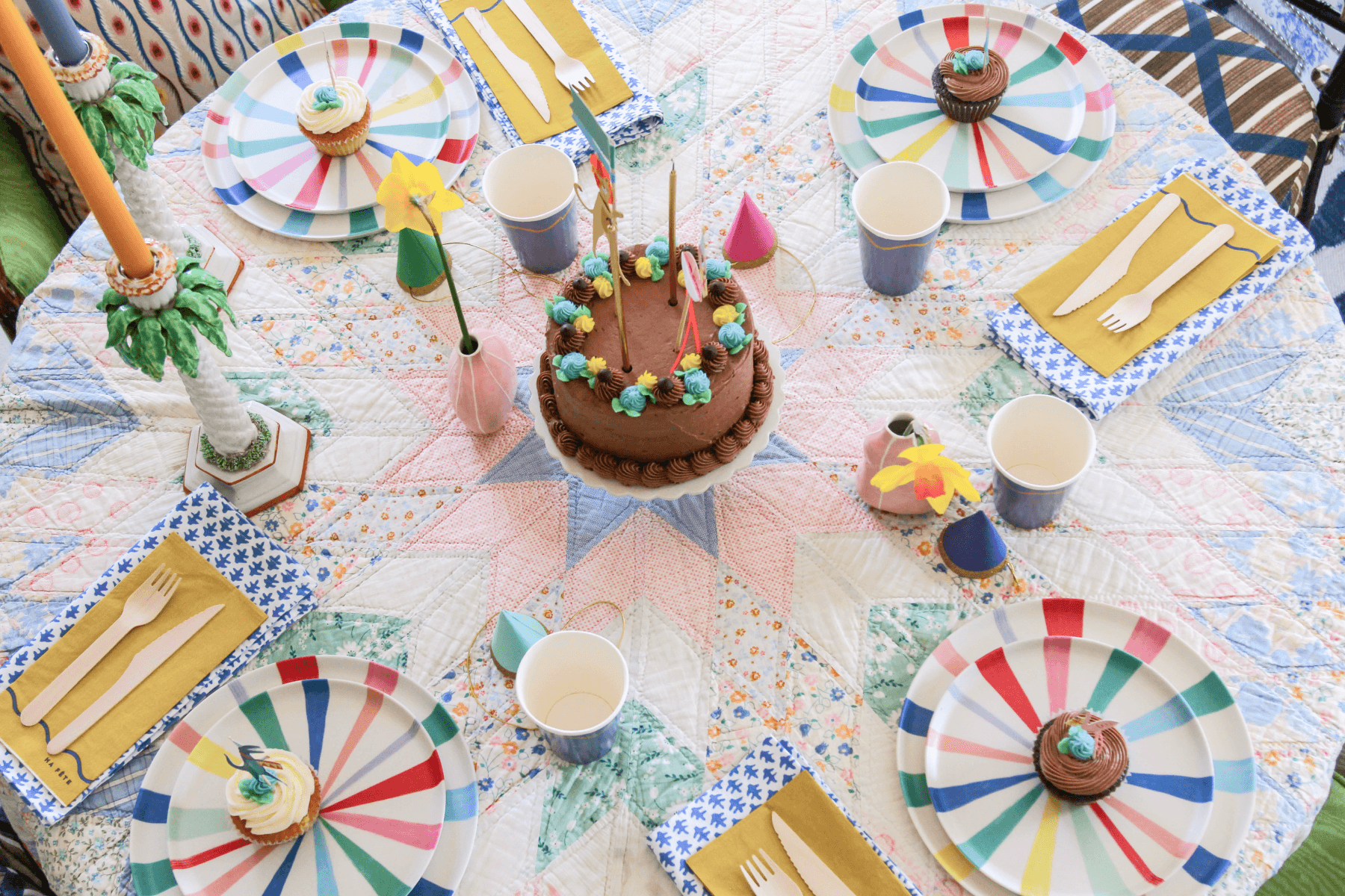 Kids' birthday party table set with a quilted tablecloth, rainbow plates, tiny party hats, and colorful cups and napkins. 