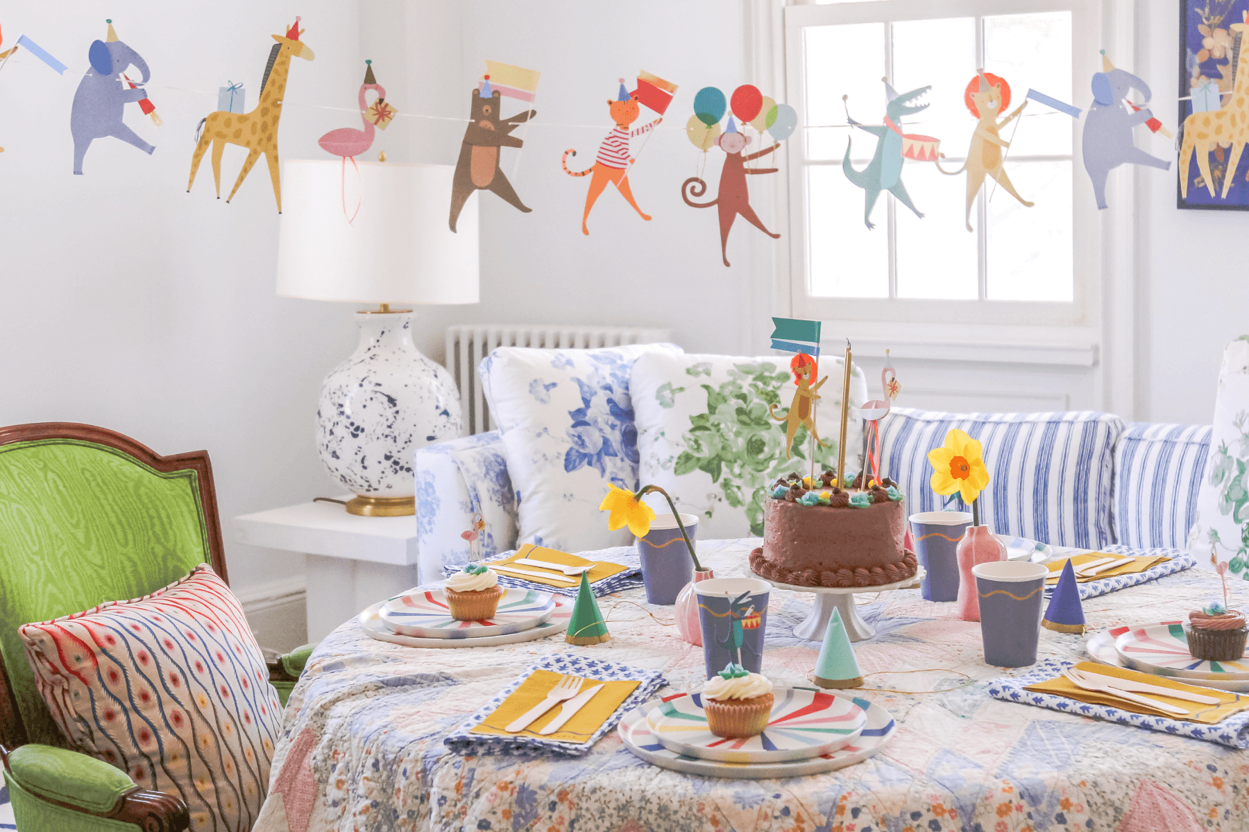 Party scene set with a quilted tablecloth, animal-themed kids' birthday decorations, and a chocolate cake.