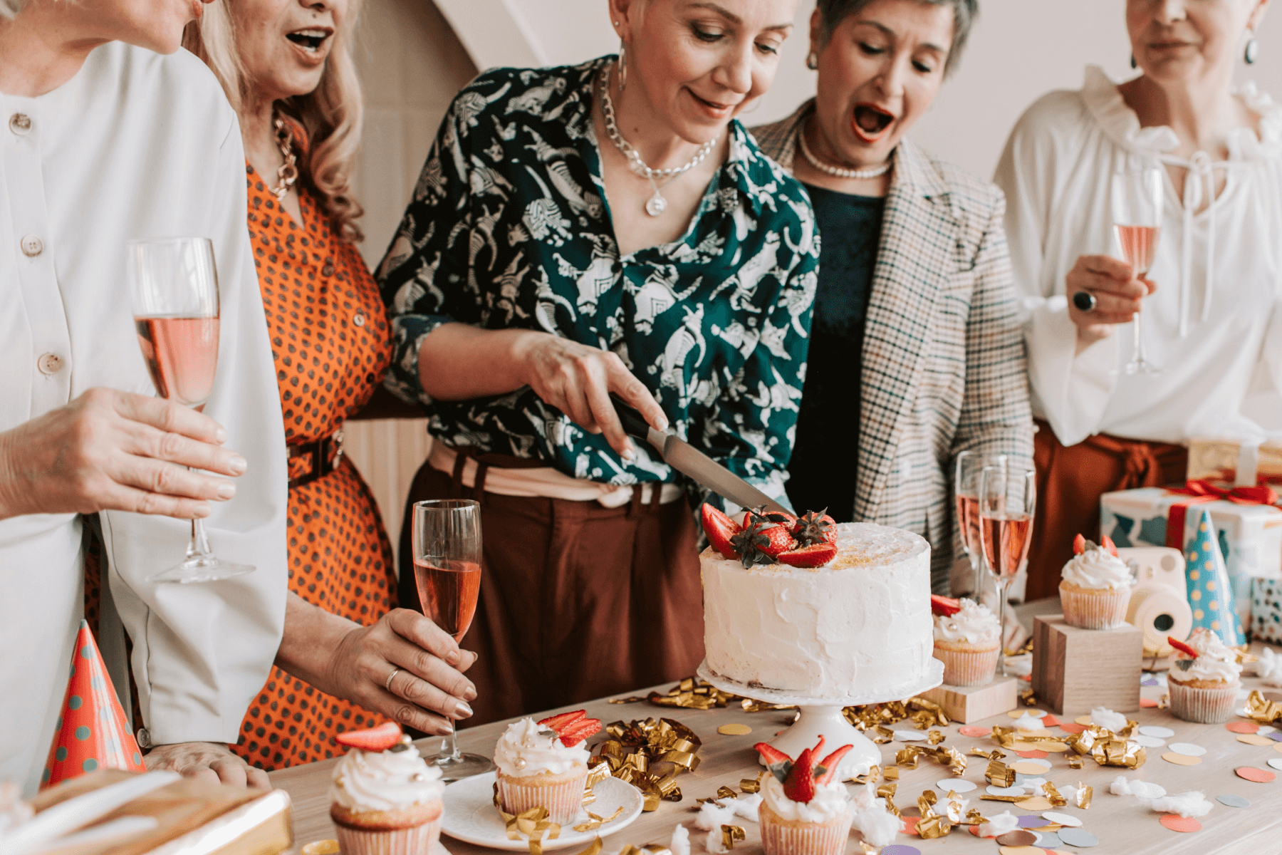 Five women gather around a table covered in cupcakes, party hats, confetti, and glasses of pink wine while one cuts into a white cake with strawberries on it.