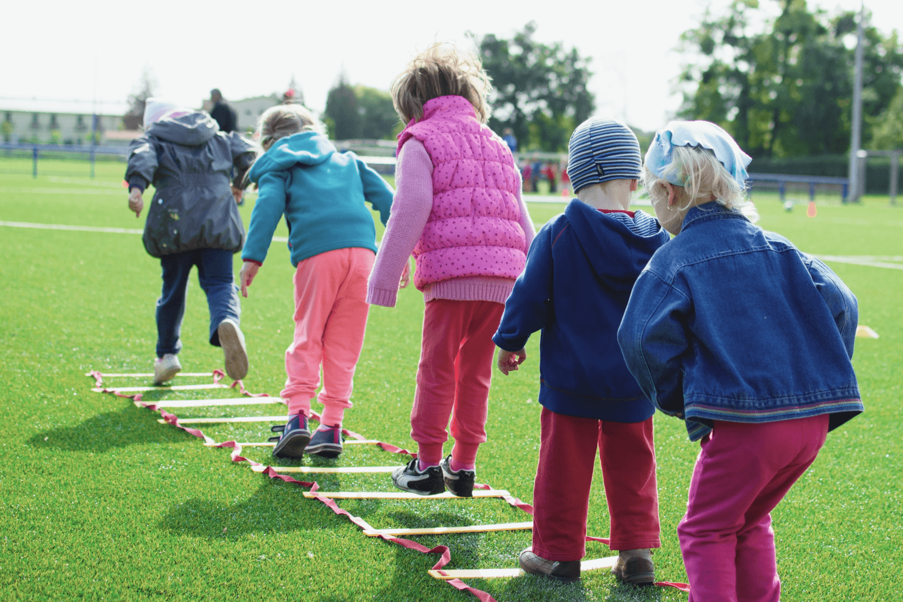 Five children are shown from behind on a green sports field, jumping through an obstacle course on the ground.