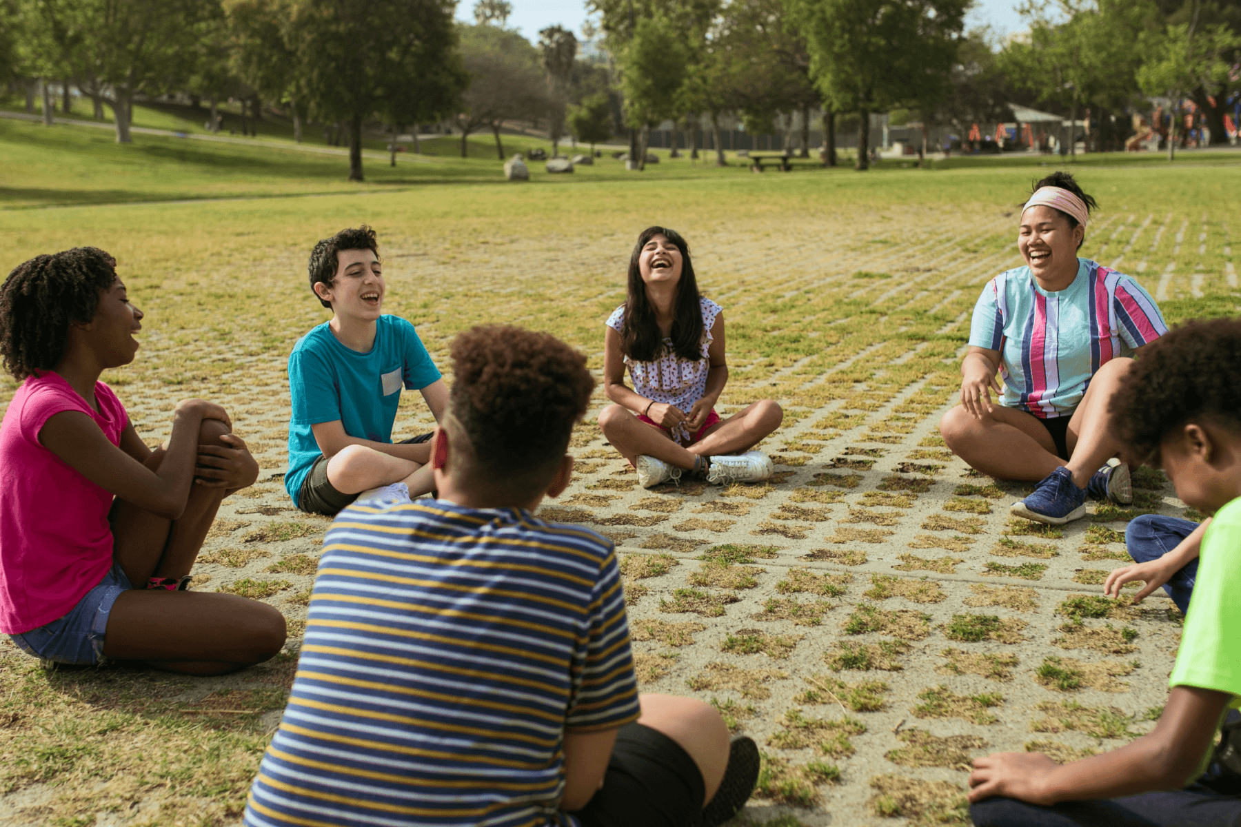 Six smiling tweens sit cross-legged in a park in a circle formation.