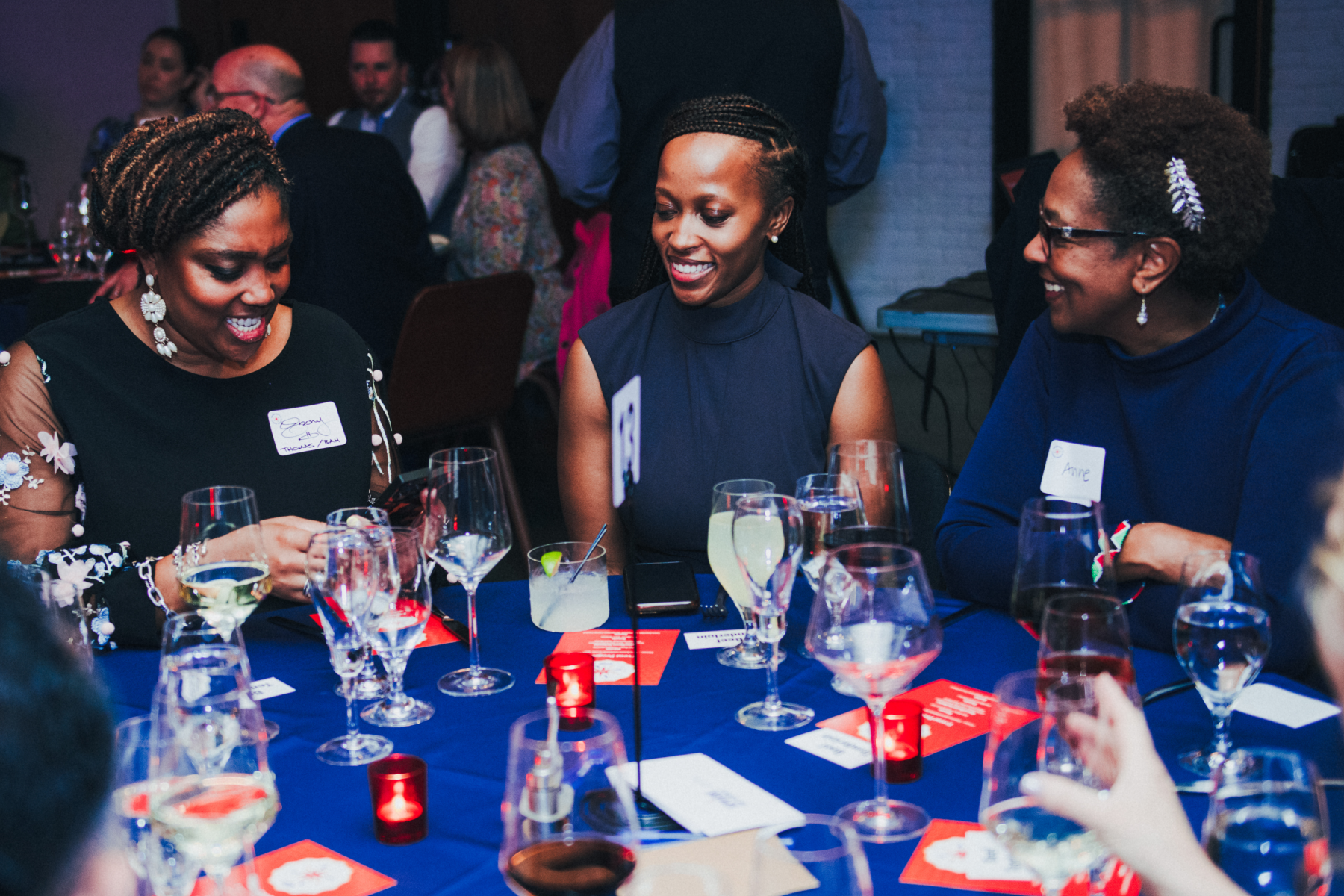 Three women wearing name tags smile as they sit around a blue table with many drinking glasses.