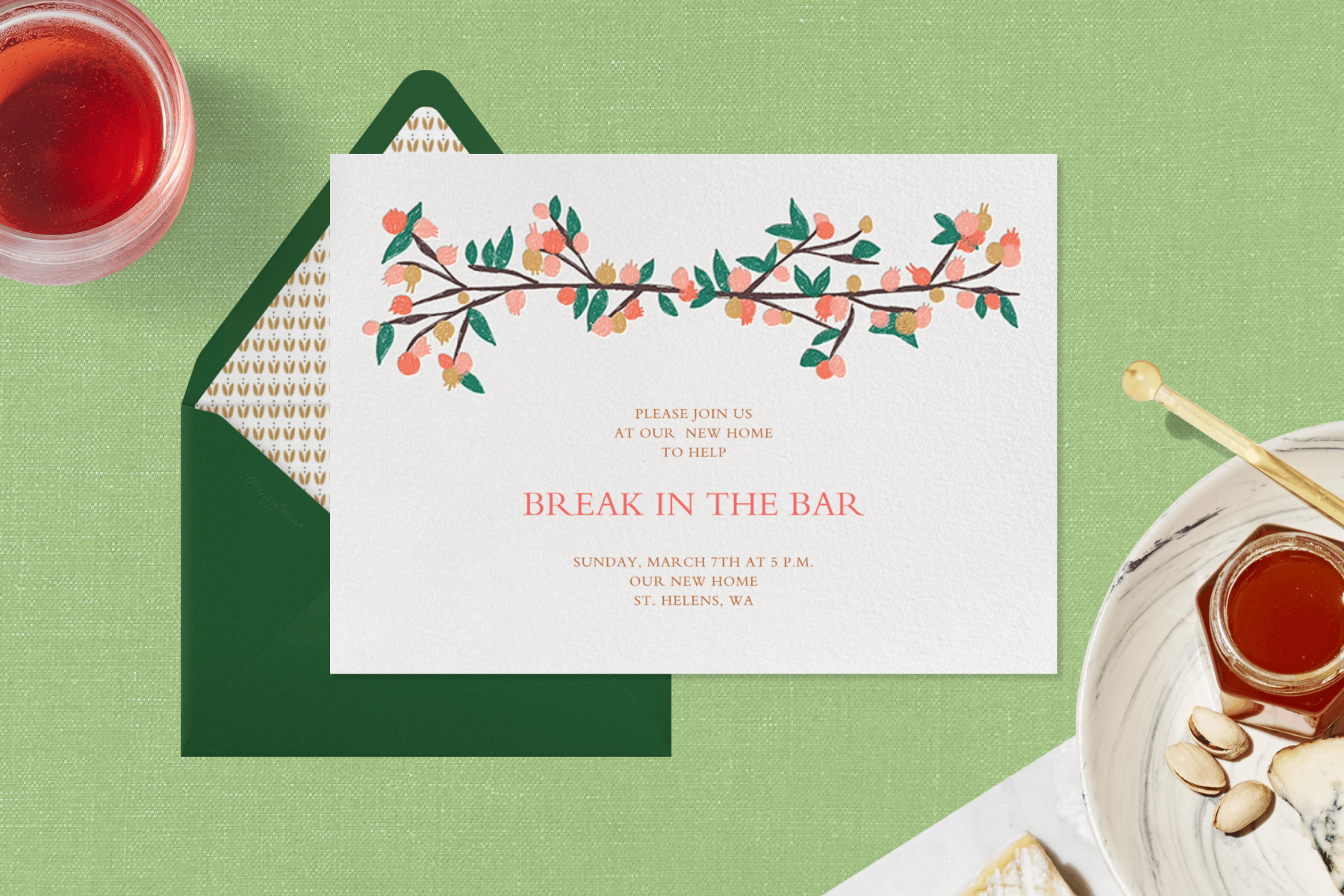 A white invitation with a branch with pink and gold berries and green leaves and the words “Break in the bar” lies on a green background with a dark green envelope, a red drink, and a plate with cheese, nuts, and honey.