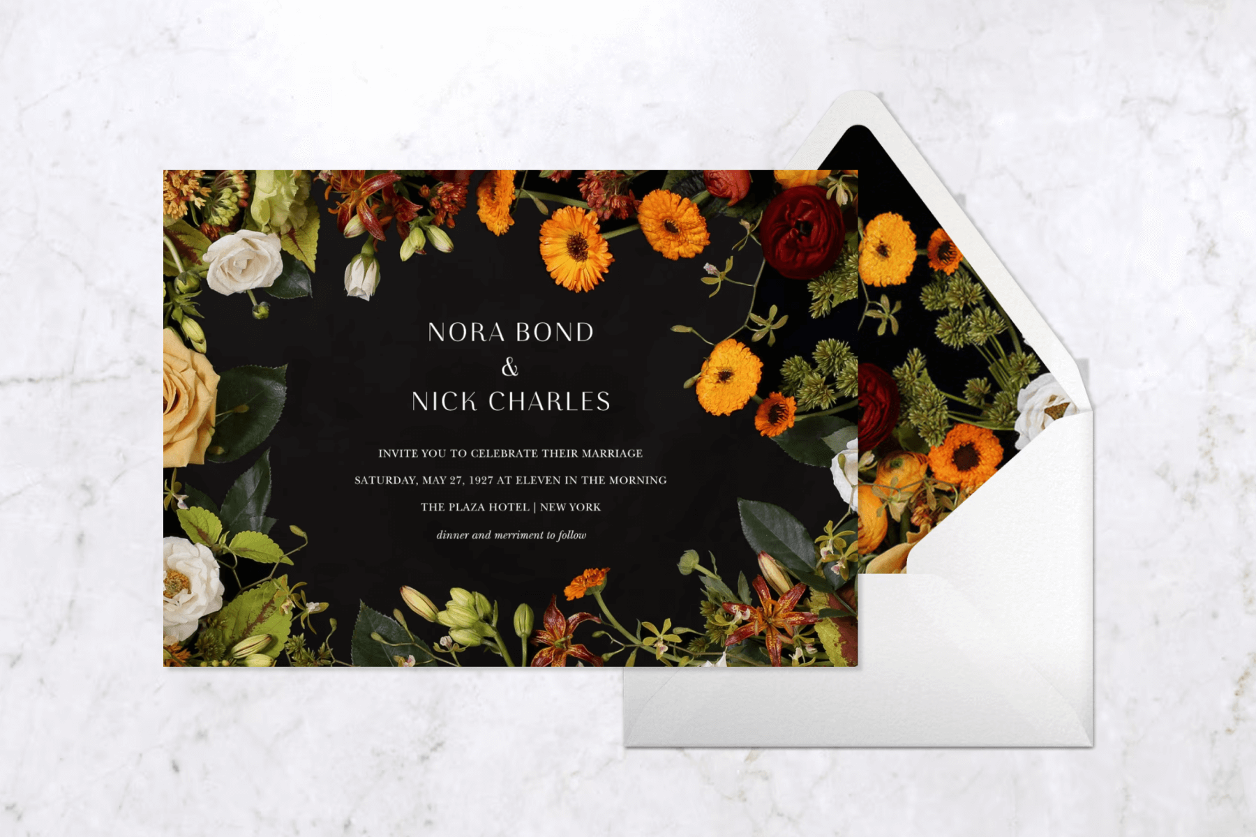 A floral wedding invitation with a black background and border of orange and cream flowers and greenery, paired with a white envelope and a matching floral envelope liner on a white marble background.