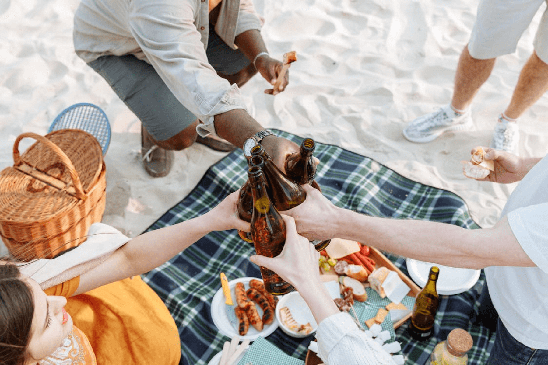 A picnic set up on a beach featuring four hands toasting beer bottles, plus a picnic blanket, picnic basket, and plates of food.