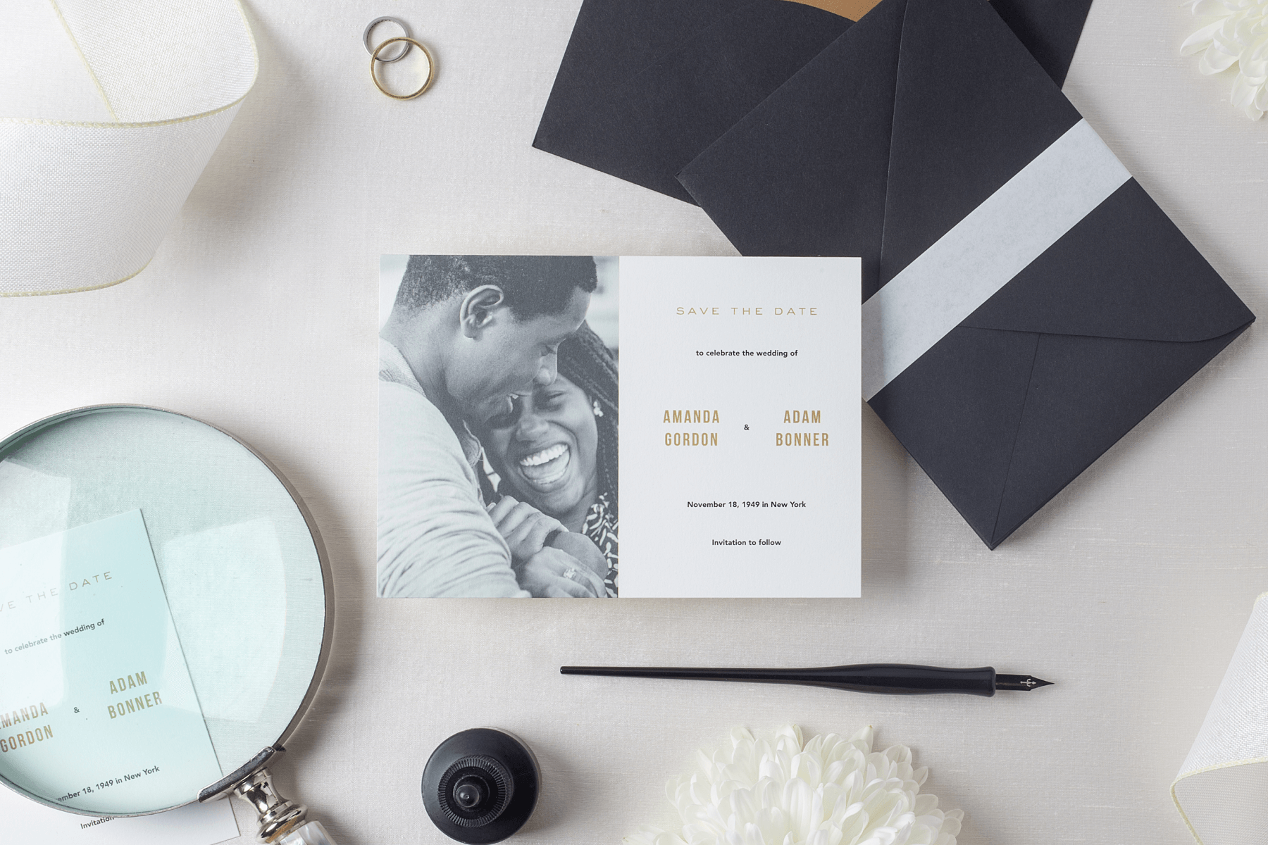 A printed save the date photo card featuring a black and white portrait of a couple with gold writing. Next to the card is a stack of black envelopes, a pair of wedding rings, a magnifying glass, flowers, and an ink bottle and pen. 