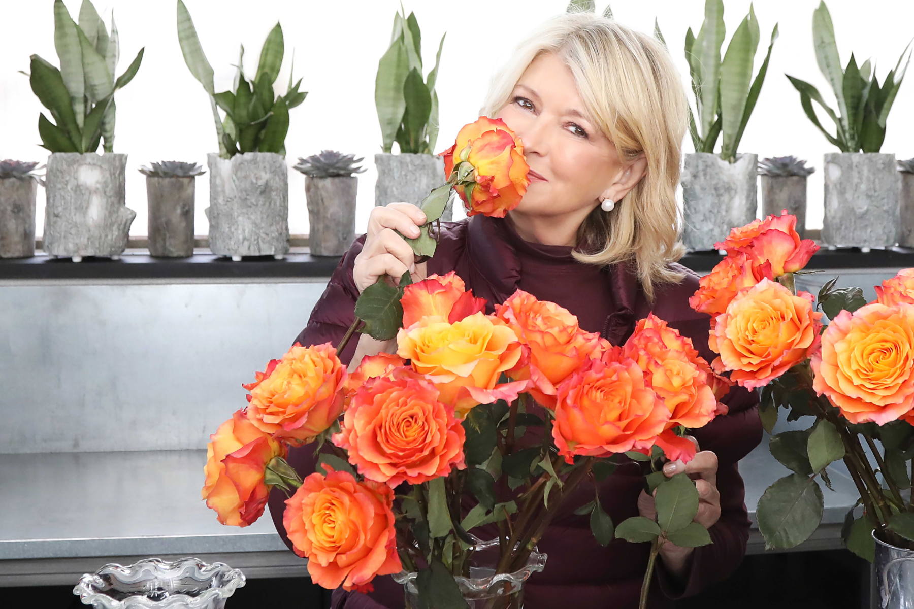 Martha Stewart stands behind a large floral arrangement of orange flowers, smelling one of them. 