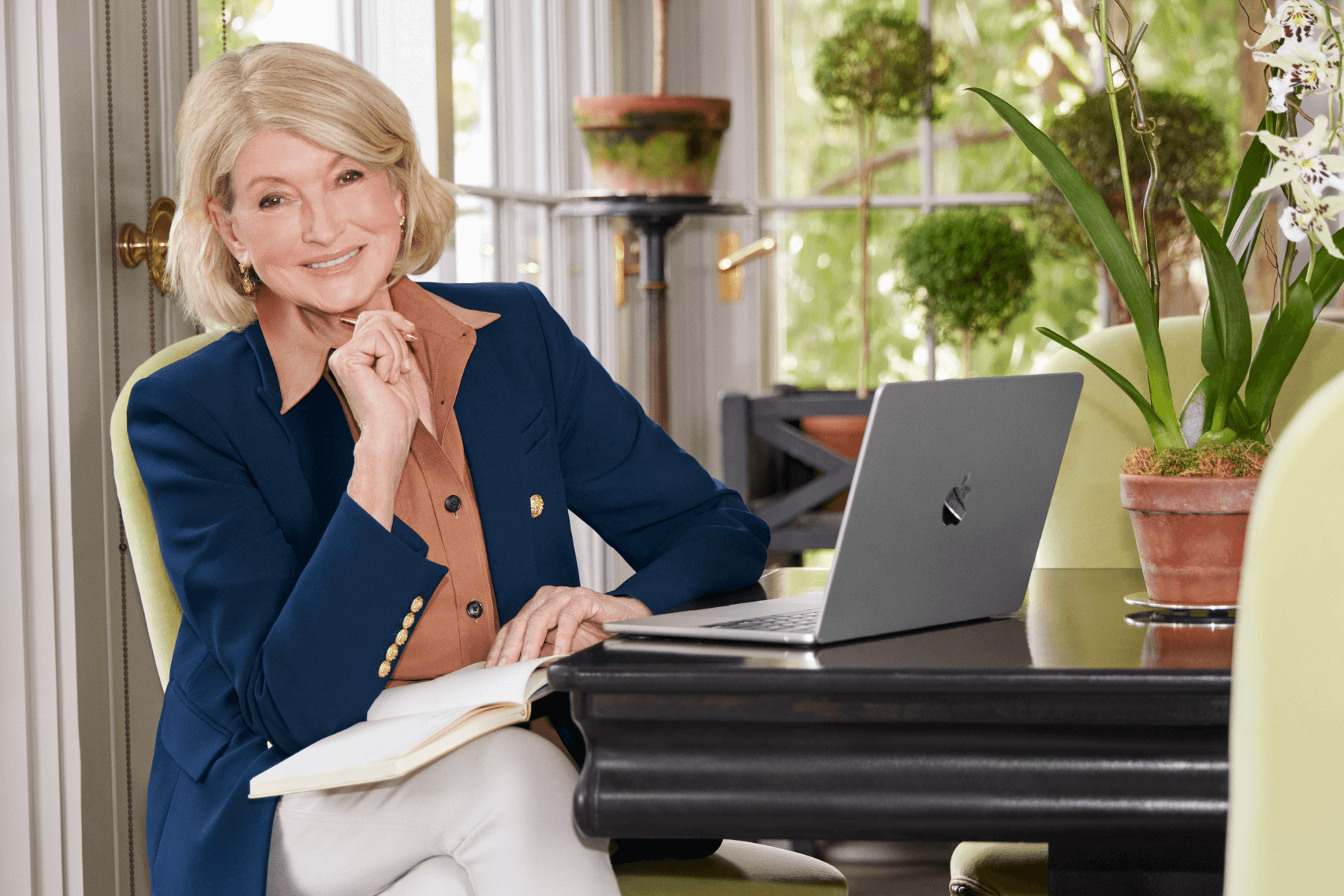 A photo of Martha Stewart sitting at a table with a journal, laptop, and pen with windows and plants in the background. 