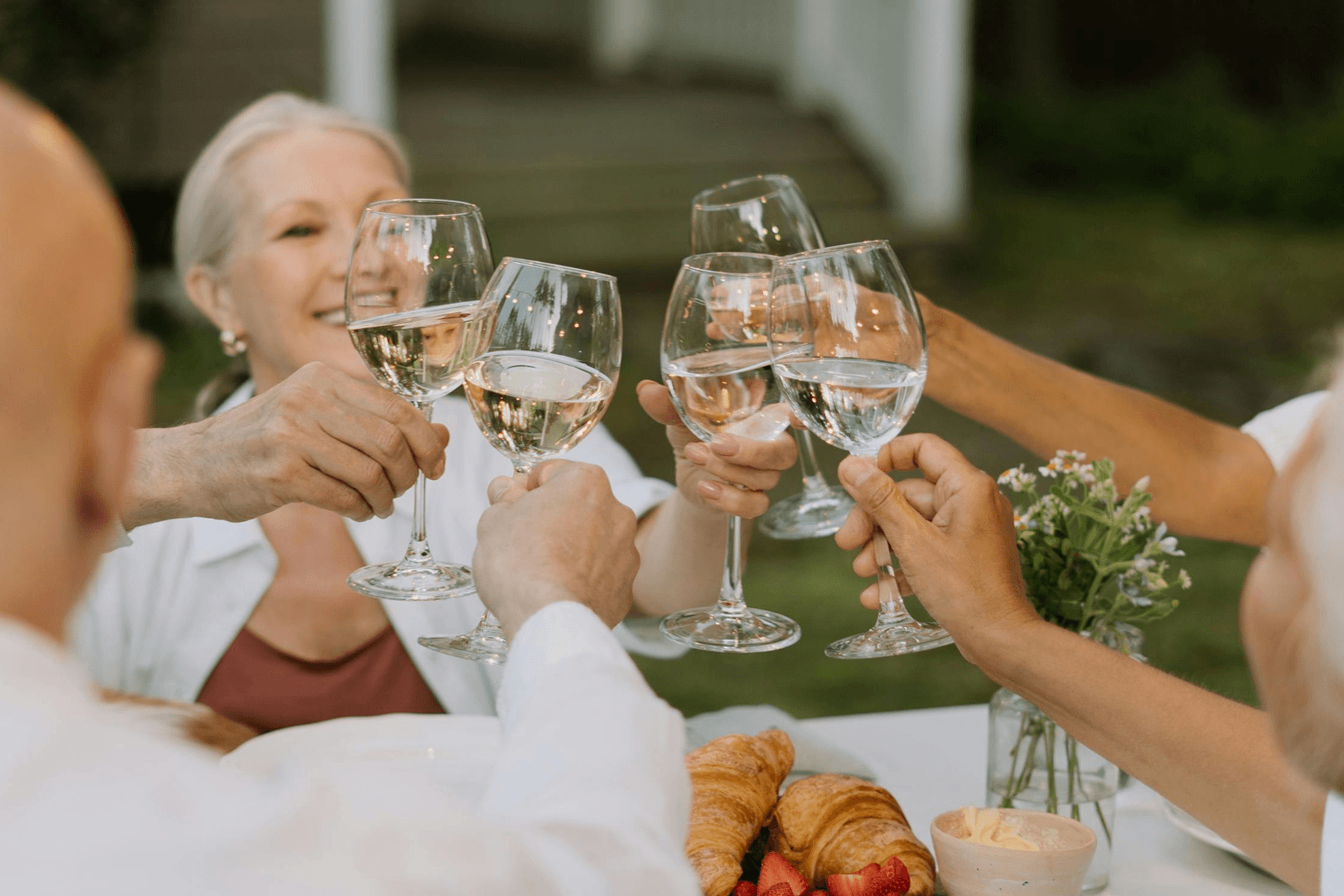ALT: Partygoers wearing white toast glasses of wine at an outdoor table topped with croissants and flowers. 