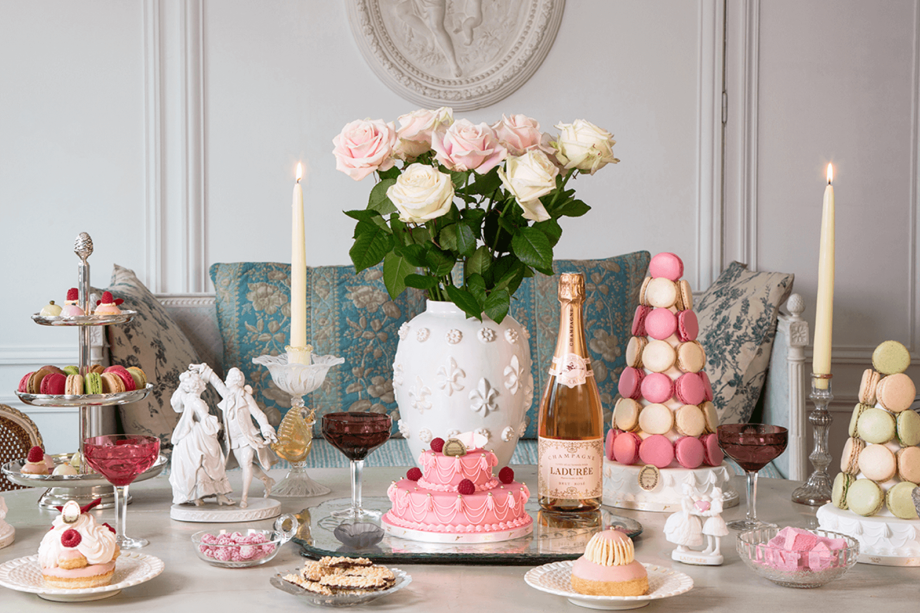 A veritable spread of delectable desserts including tiers of macarons, a cake, and other pastries along with rosé wine, roses, and candles on a bare white table.
