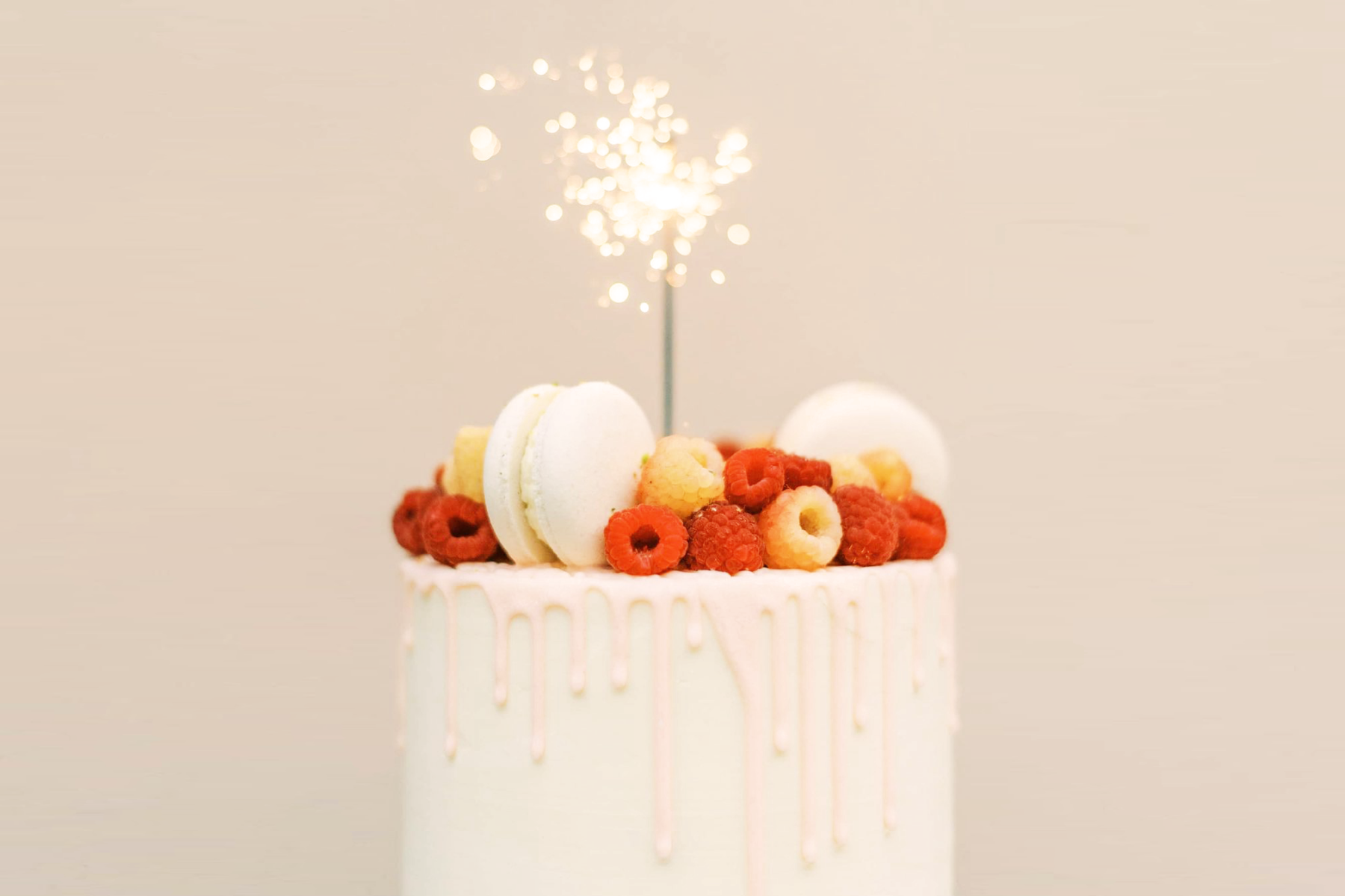 A cream-colored birthday cake with macarons, raspberries, and a sparkler candle.