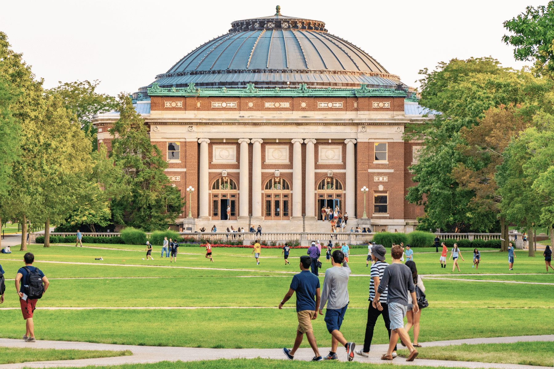 Students walk across a green lawn in front of a domed brick building on a college campus.