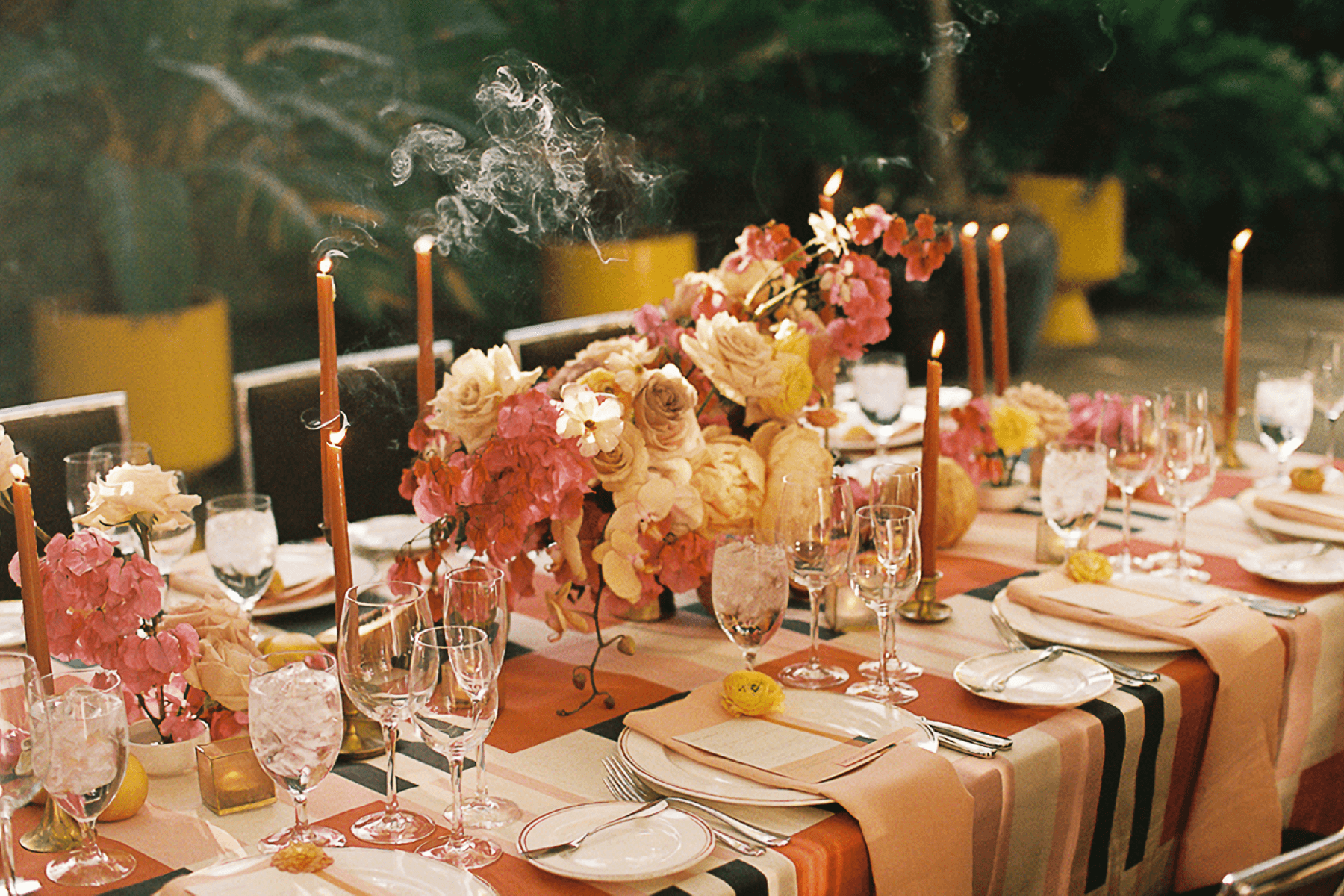 A crowded outdoor banquet table is set with flowers, candles, and a striped tablecloth in shades of pink, yellow, and orange.