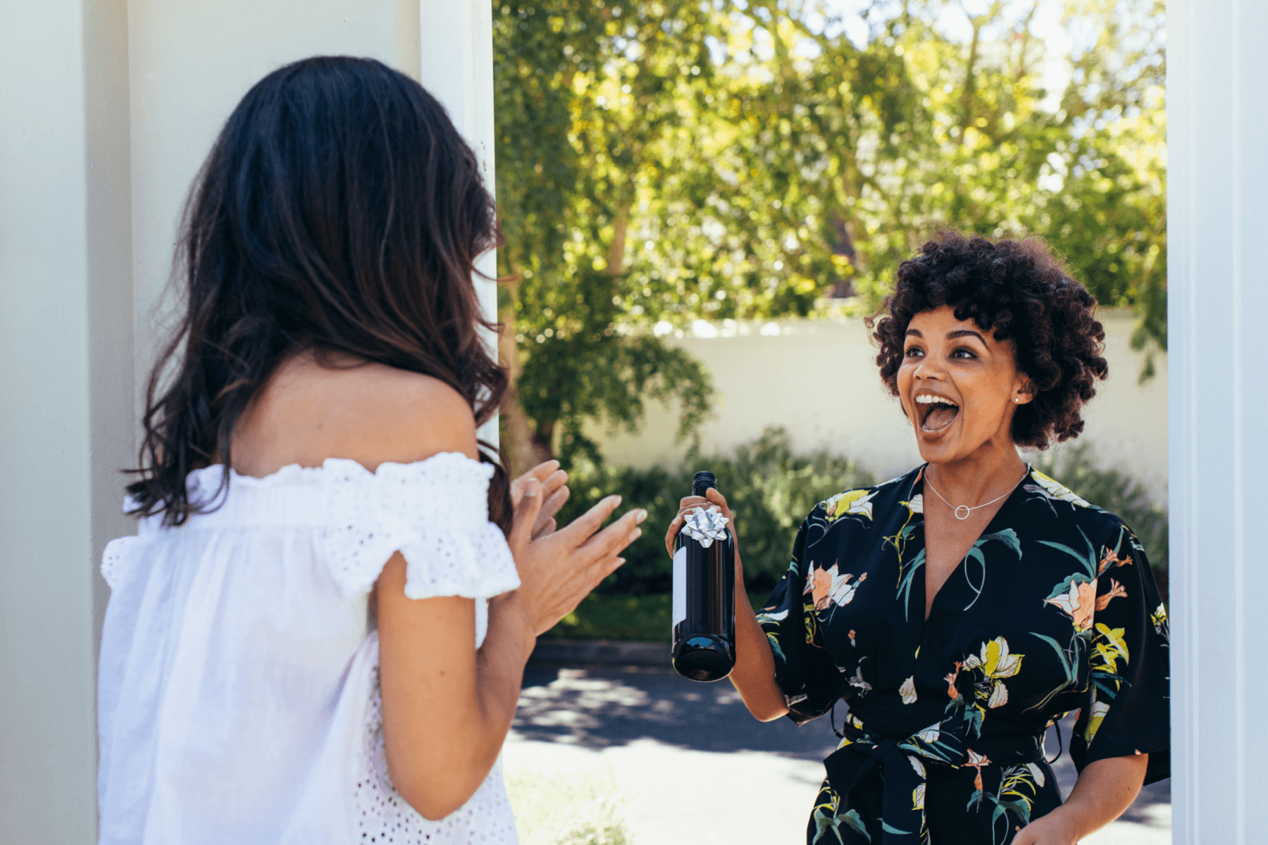 A woman greets another woman, smiling, at the door with a bottle of wine with a silver bow on it.