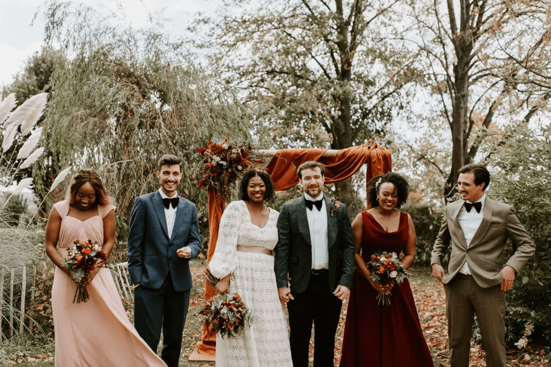 men and women wearing dressy outfits stand at a rustic outdoor wedding altar.