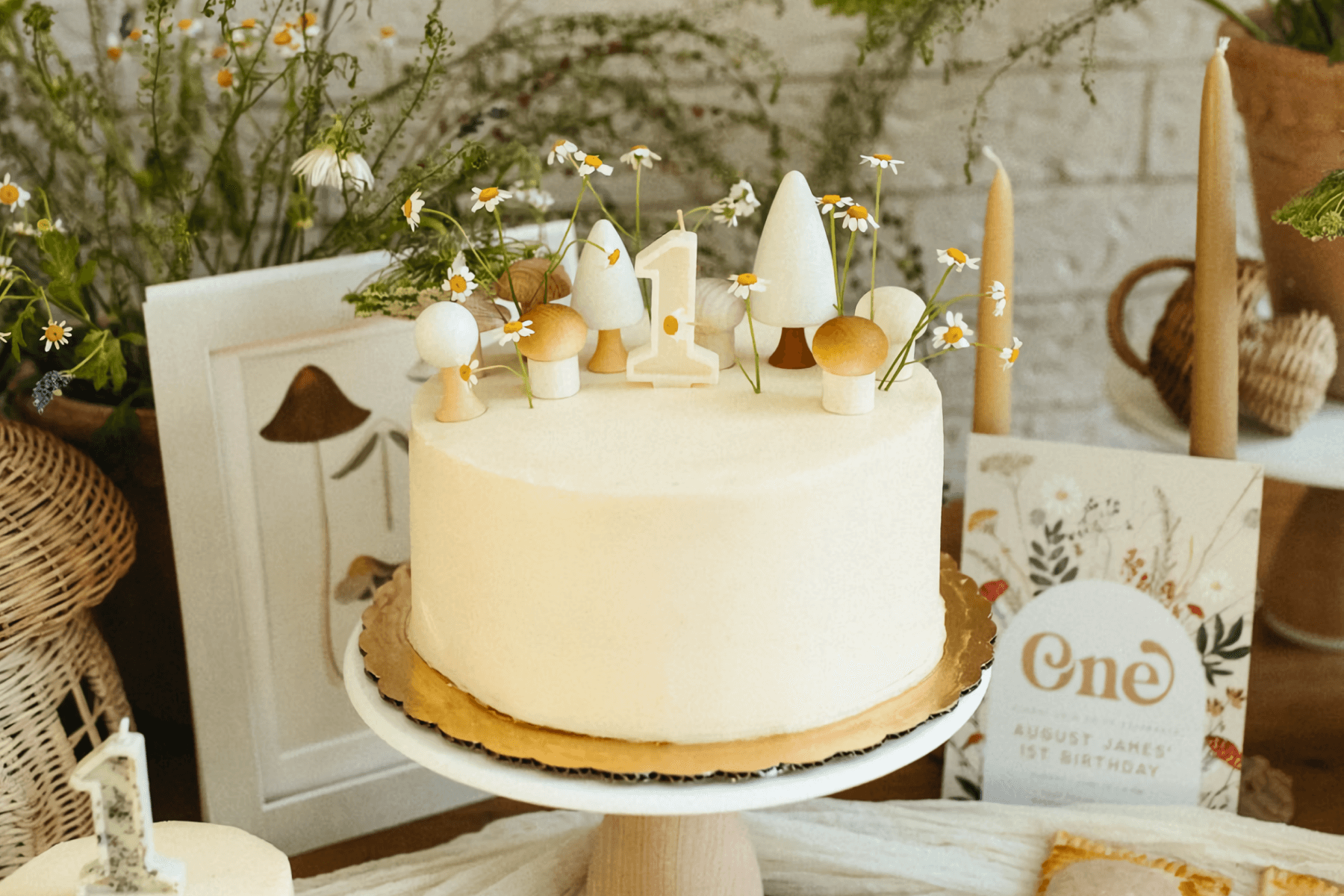 A white layer cake on a crowded table is decorated with mushrooms, chamomile flowers, and a white “1” candle.
