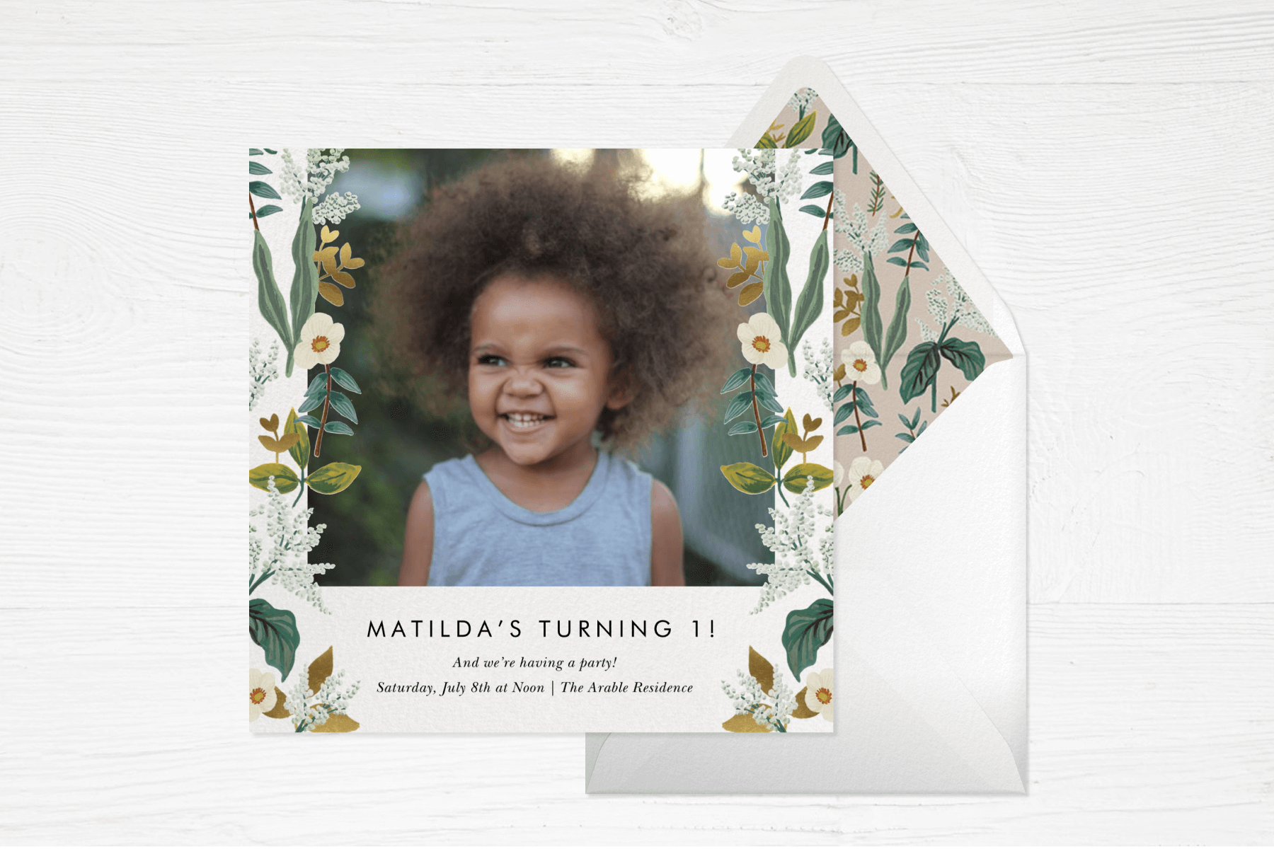 A birthday invitation reads “Matilda’s turning 1!” with a photo of a young girl and a leafy floral border on each side.