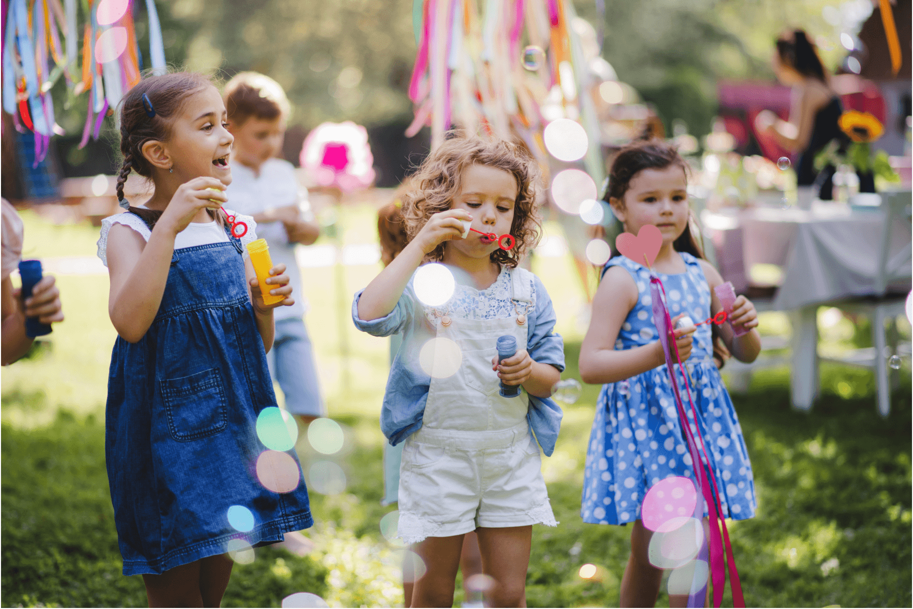 Three young children blow bubbles at an outdoor party with streamers behind them.