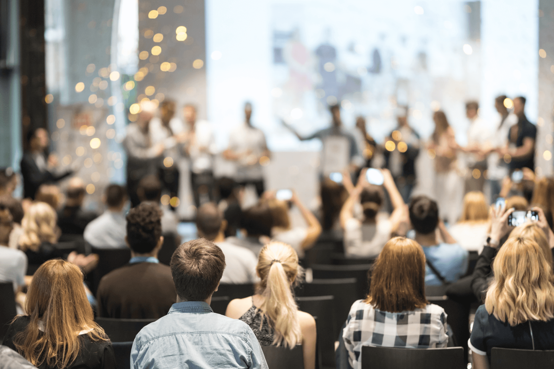 A group of people in an auditorium is viewed from the back with people onstage blurred.