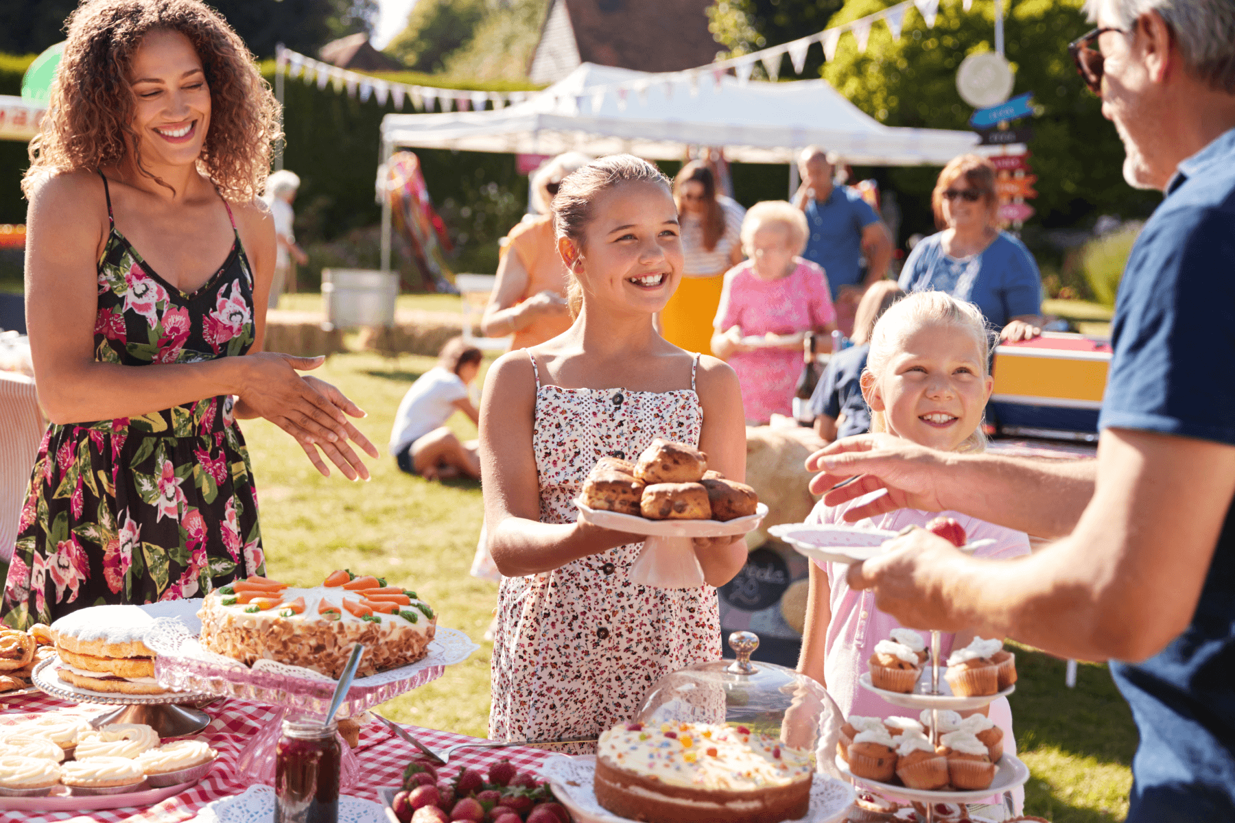 Two young girls at a dessert table in a park hold out baked goods for a man to take while a smiling woman looks on.