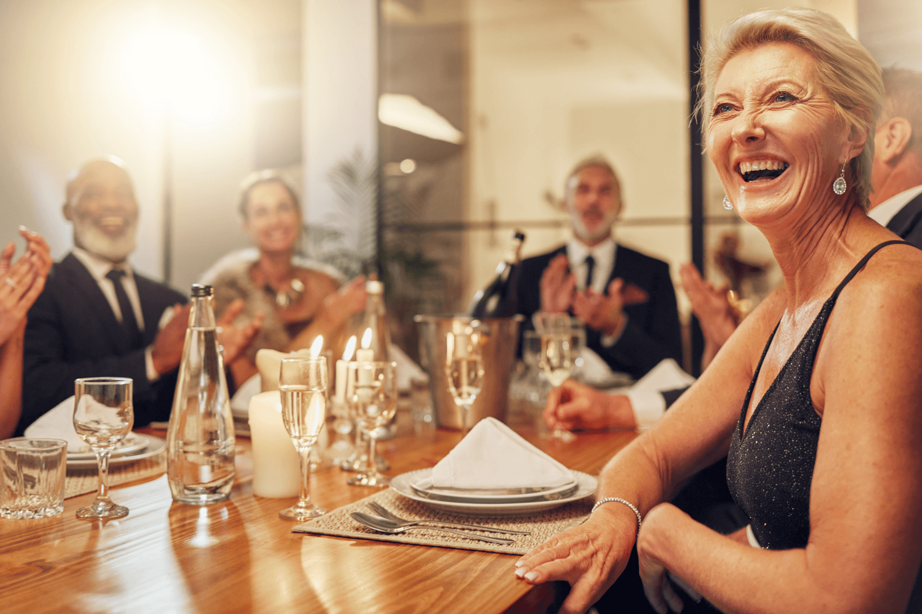 A laughing woman at a dinner table with other well-dressed guests, who are clapping.