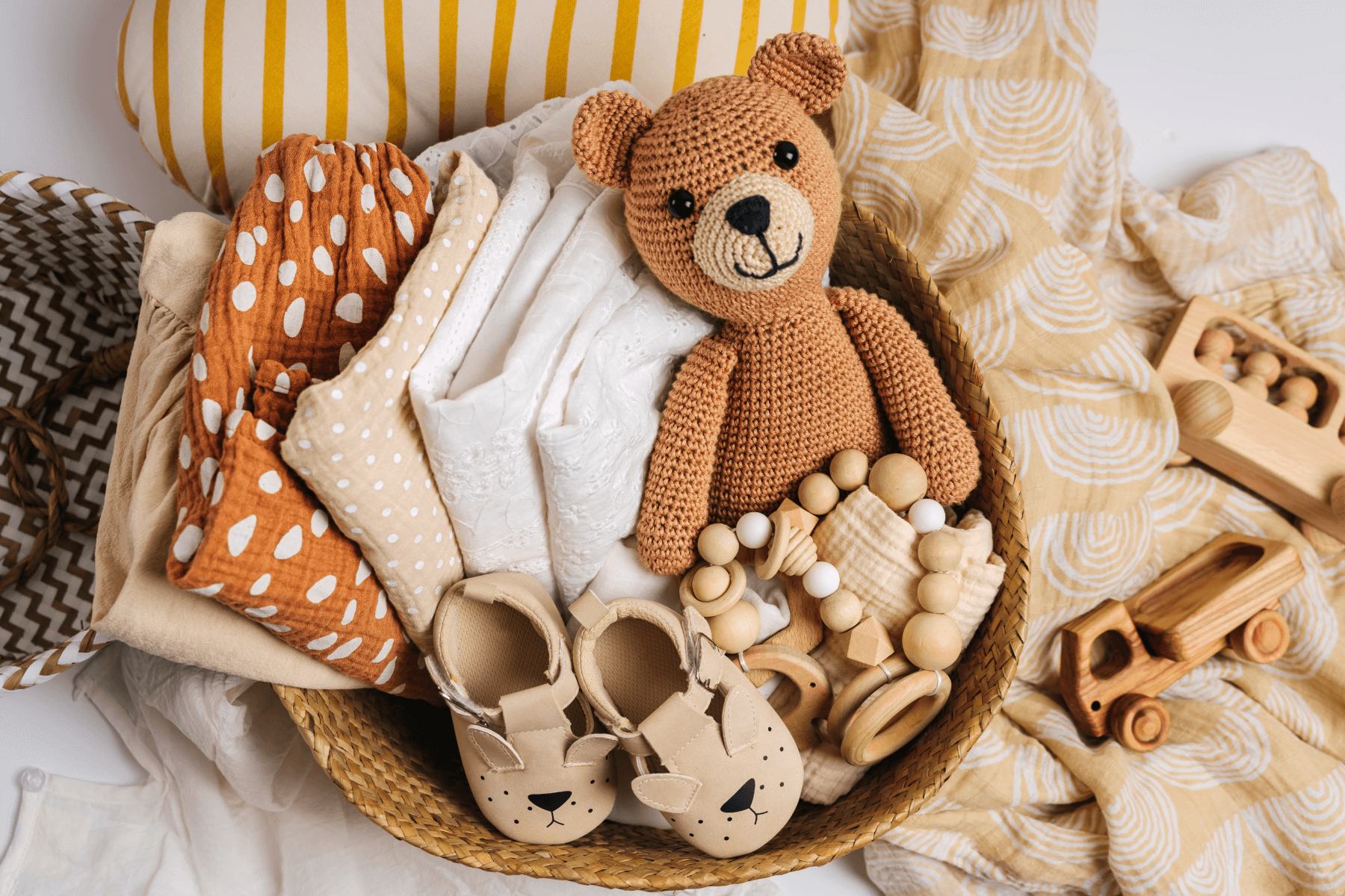 A woven basket shown from above is filled with neutral colored and polka dotted linens, a knit teddy bear, wooden baby teething toys, and baby shoes.