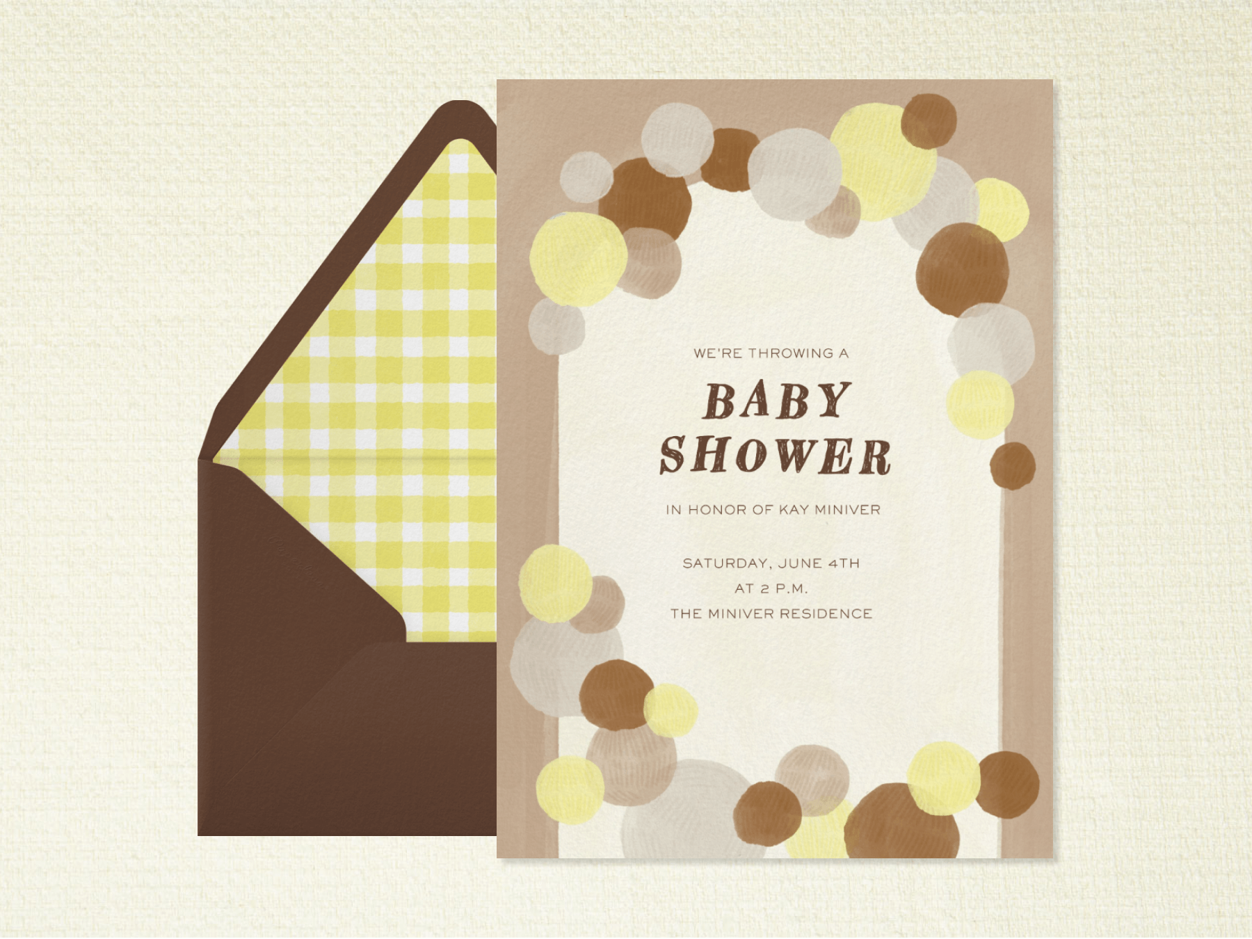A baby shower invitation with brown, gray, and yellow circles representing balloons form an arch around the party details, beside a brown envelope with lime green gingham liner.