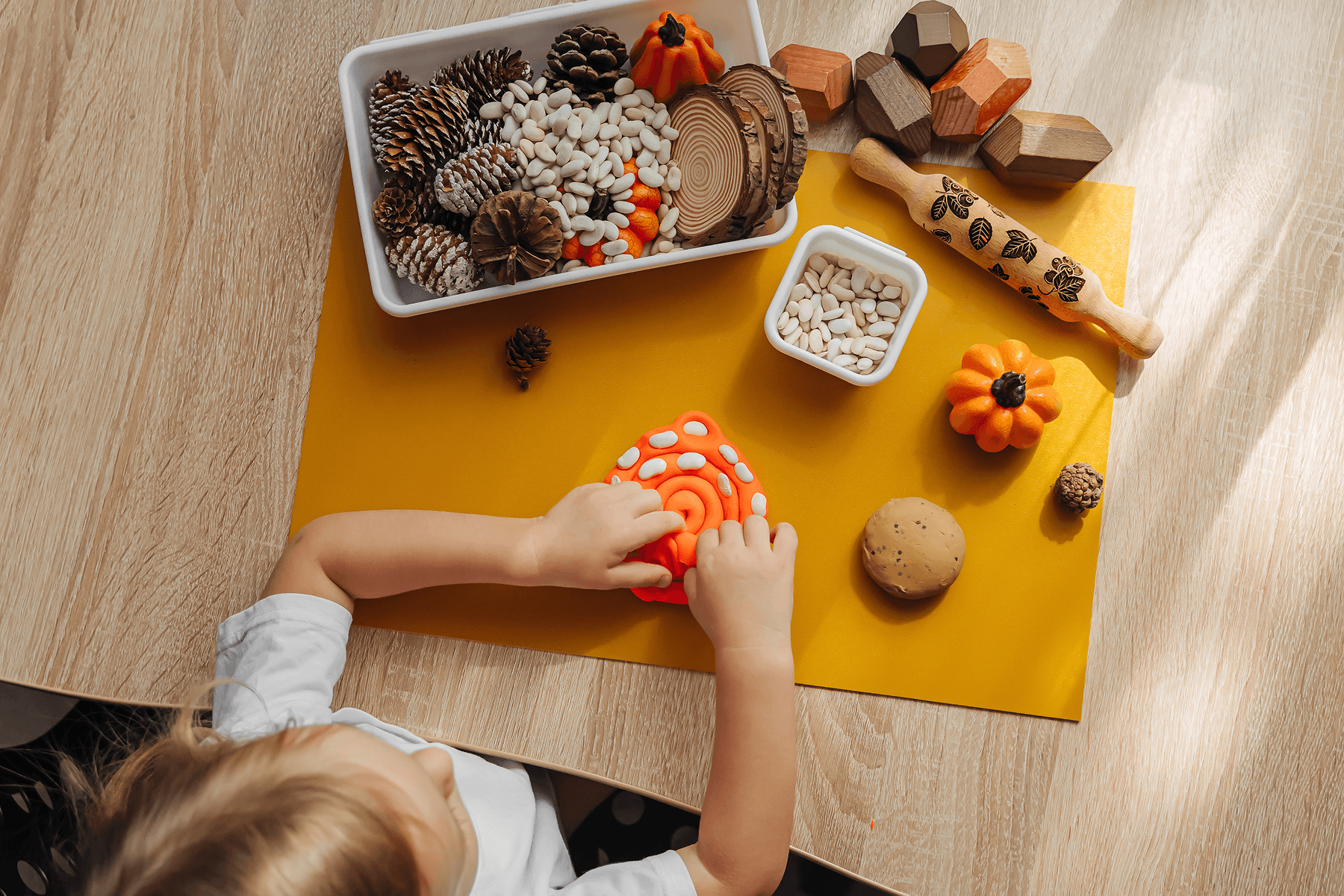 ALT: Seen from above, a child presses white beans into orange play dough on a yellow mat, surrounded by a bin of pinecones, beans, and wood slices, small pumpkins, a patterned rolling pin, and wooden blocks.