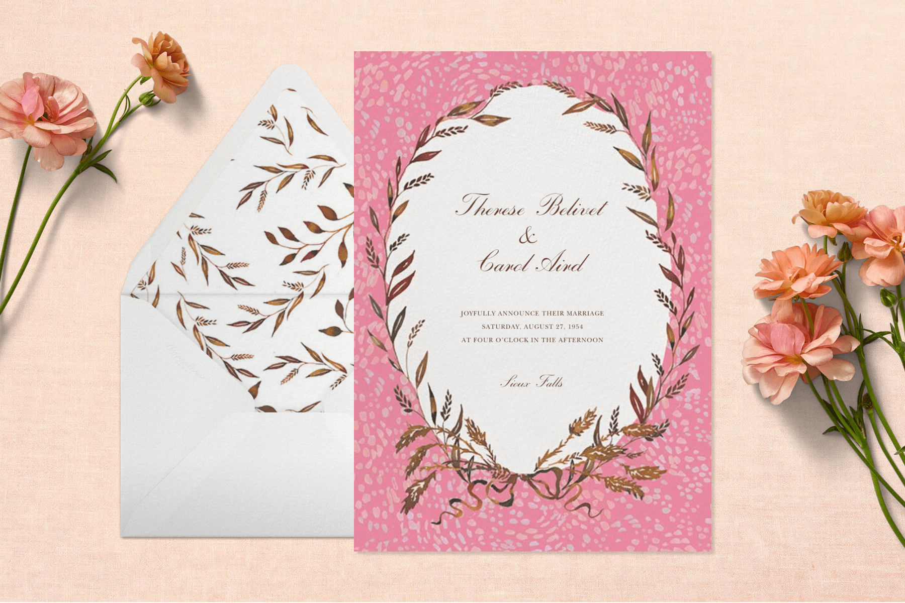 On a light pink backdrop adorned with flowers sits a pink and white wedding invitation with gold floral details next to a white envelope with floral lining.