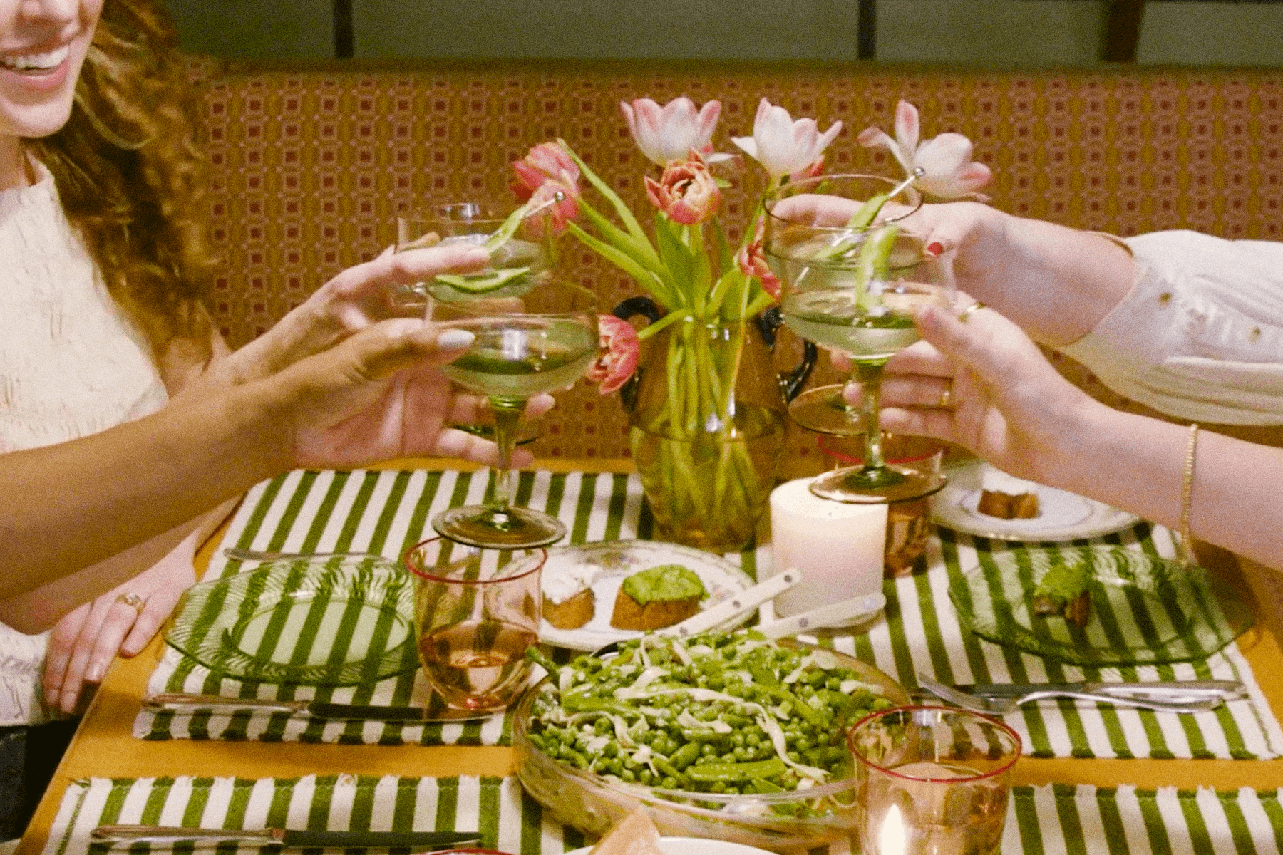 ALT: Four hands toast martini glasses with snap pea garnishes over a dining table set with green and white striped place mats, a bowl of pasta with peas, and a vase of pink flowers.