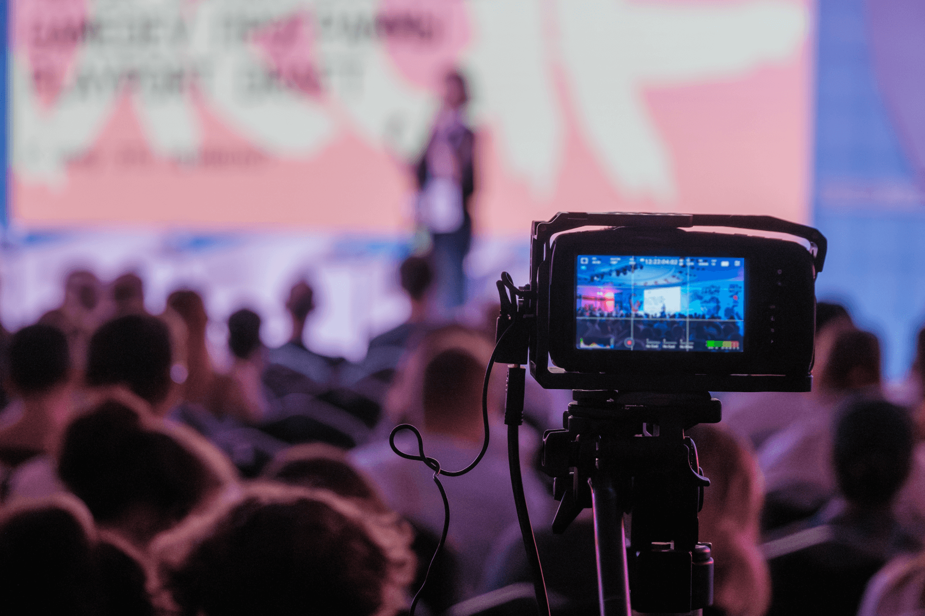 A video camera recording footage from the audience of an event with a speaker on a stage. 