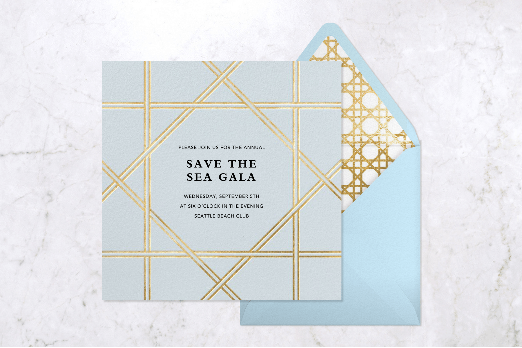 A powder-blue sea-themed gala invitation with gilded accents sits to the left of a light blue envelope with a golden-stitched interior, against a white marble backdrop.