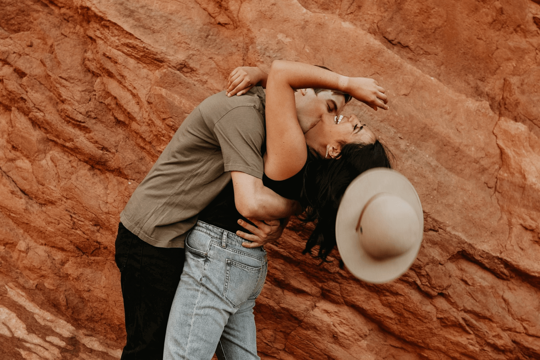 A man kisses a woman whose hat is falling off as they bend playfully in front of a red rock