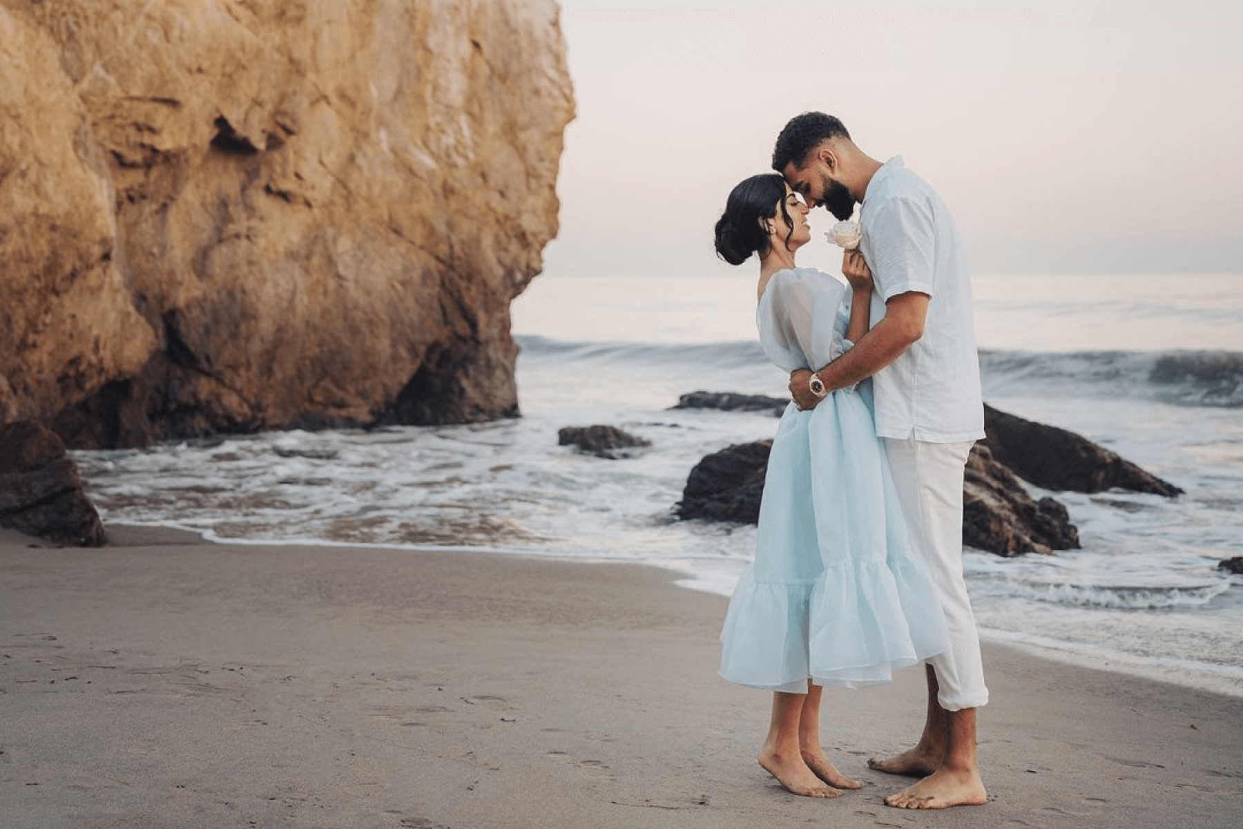 man and woman embrace on the shore with large rocks in the water