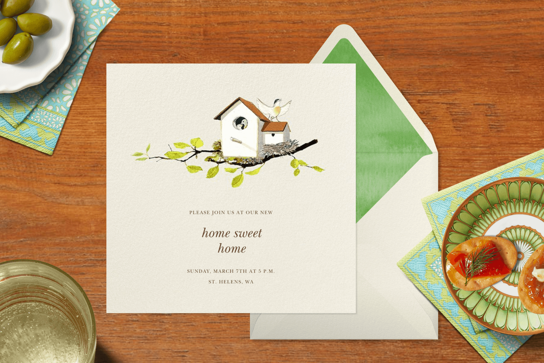 A square housewarming party invitation reads “home sweet home” with a watercolor painting of birds in a birdhouse on a branch with another bird spreading its wings on the roof. Around the card on a wood backdrop are small plates with hors d'oeuvres, a bottle of wine, and a drinking glass