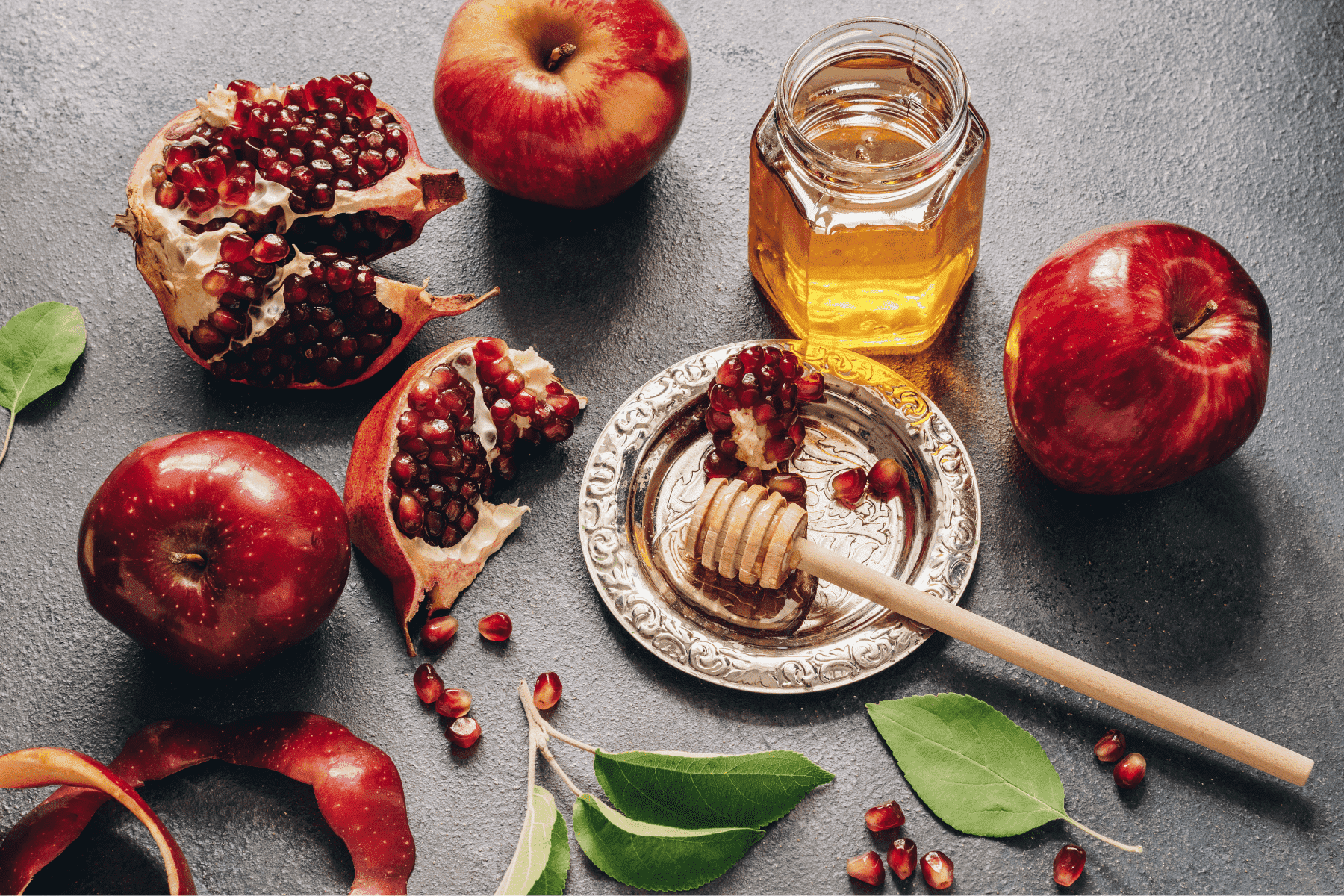 Pomegranates, apples, and a jar of honey on a surface.