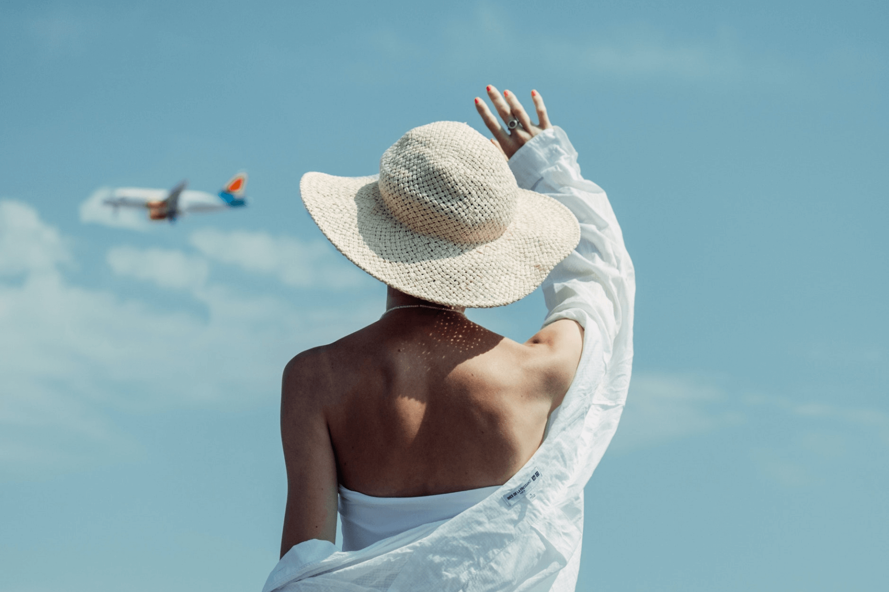 Shown from behind, a woman in a woven sun hat with a white shirt draped off her shoulder waves at an airplane in a blue sky.