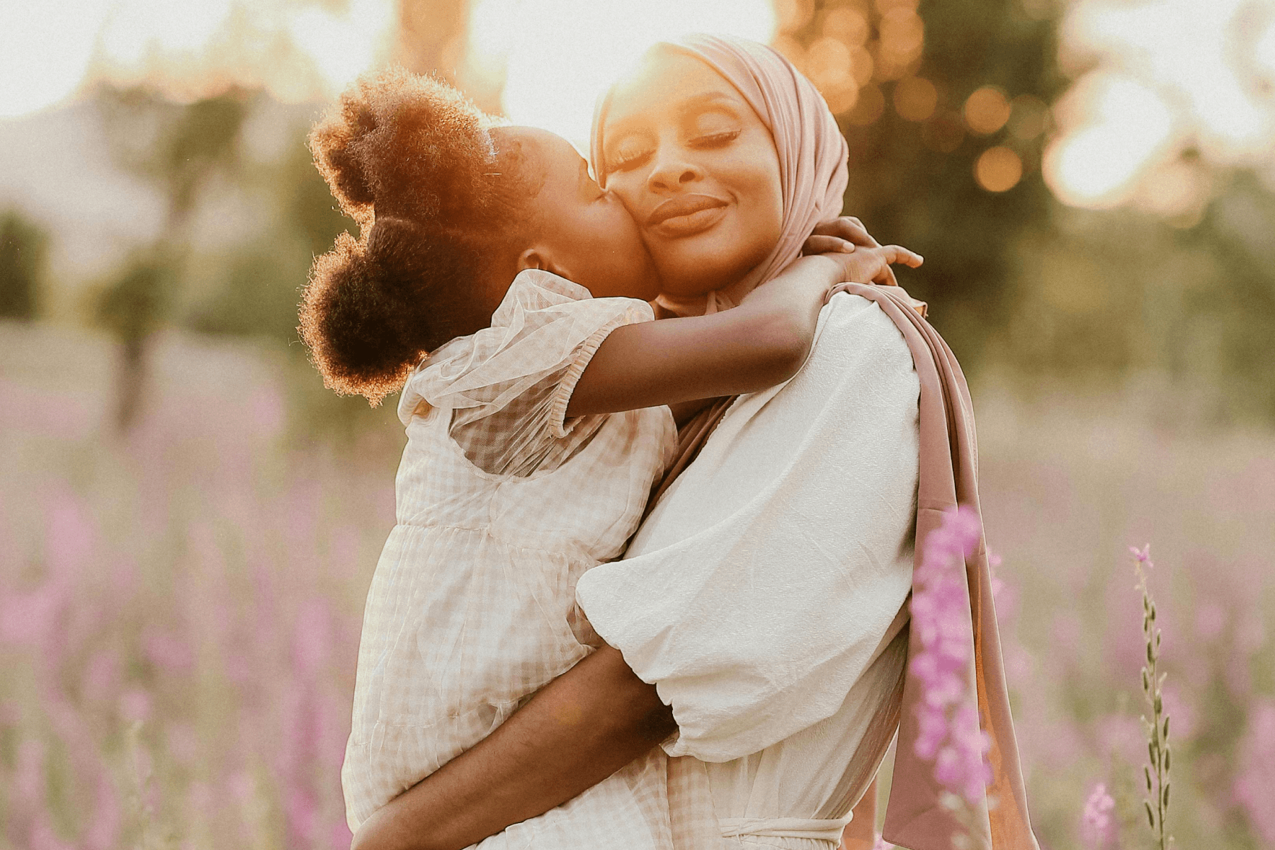 A woman stands in a field of flowers, holding up a small girl who is kissing her on the cheek.