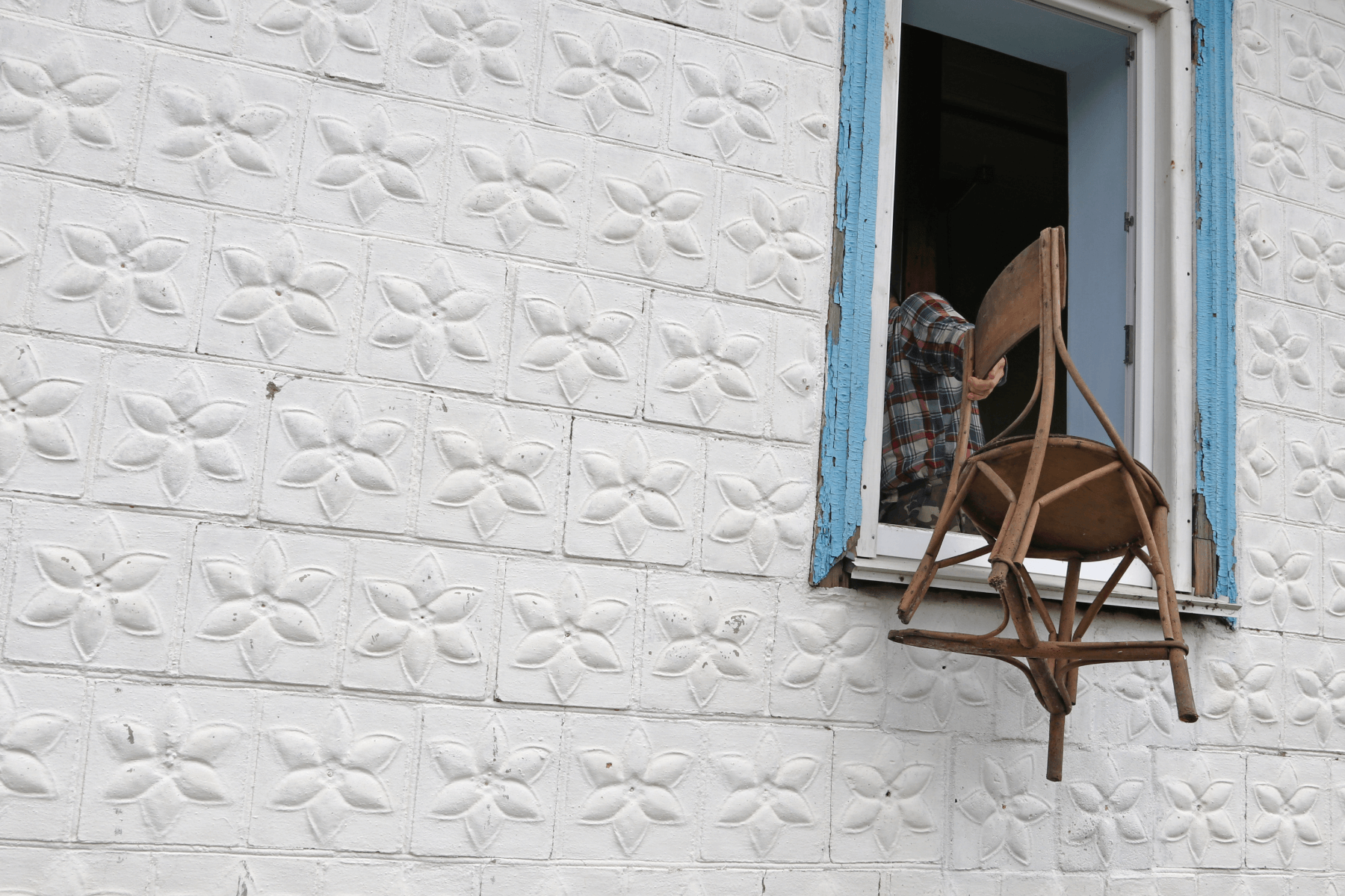 A white mosaic wall with a man hanging a chair out a window.