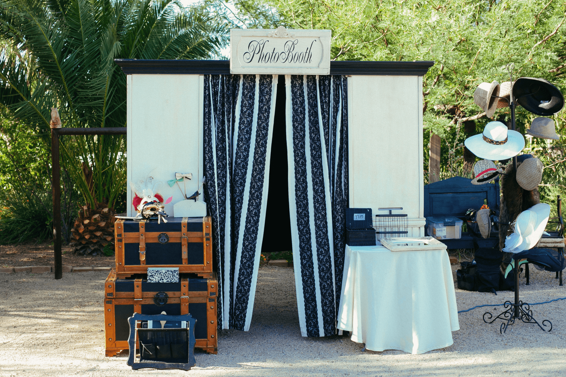 A photobooth set up outdoors with navy and white striped curtains, black and brown trunks topped with props, and a hat rack full of hats.