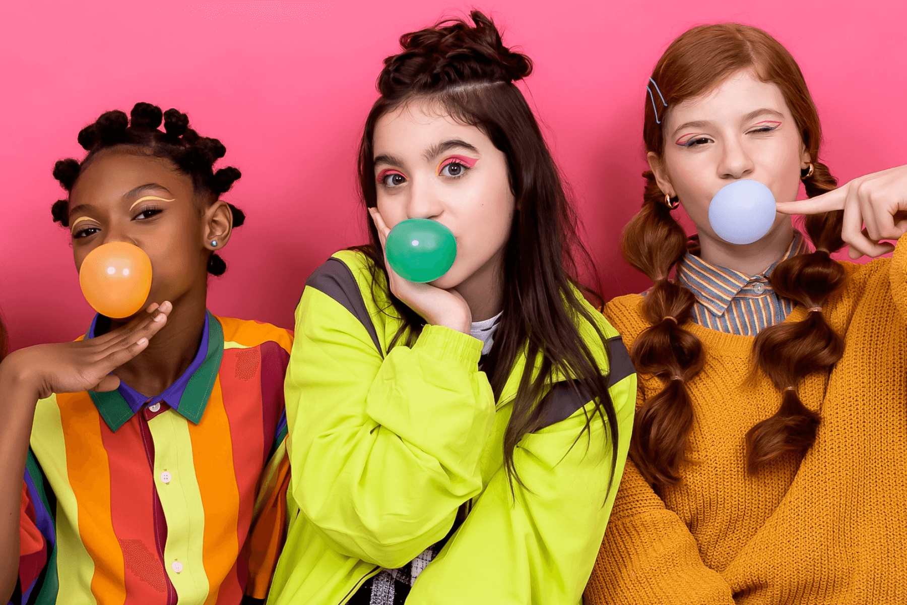 Three teenage girls in bright clothing against a pink background. They are all blowing bubbles with bright-colored gum (orange, green, and blue).