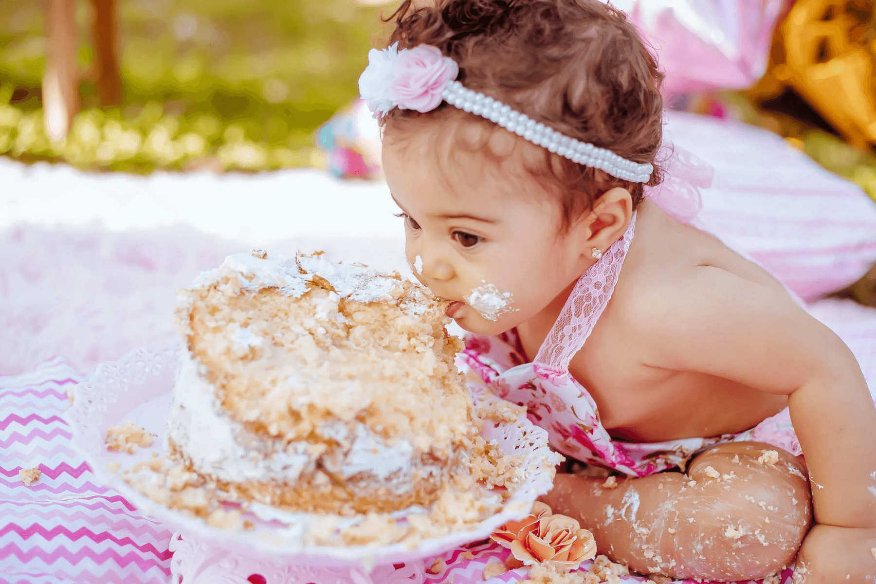A baby with a pearl headband with a pink flower, eating a birthday cake, cheeks and legs covered in icing, on a pink and white striped picnic blanket outdoors. 