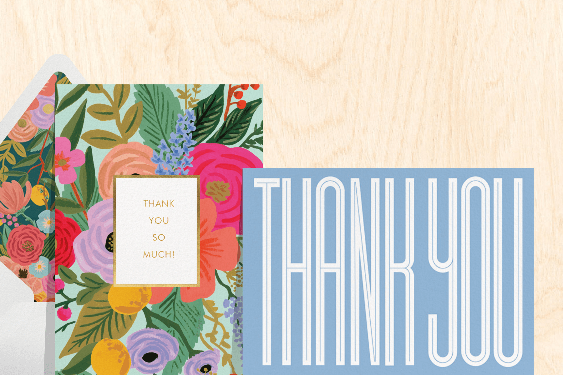A light wood background featuring two thank you cards: The first with gold text, colorful floral illustrations, and a matching envelope liner and white envelope; the second with a blue background and modern white text.