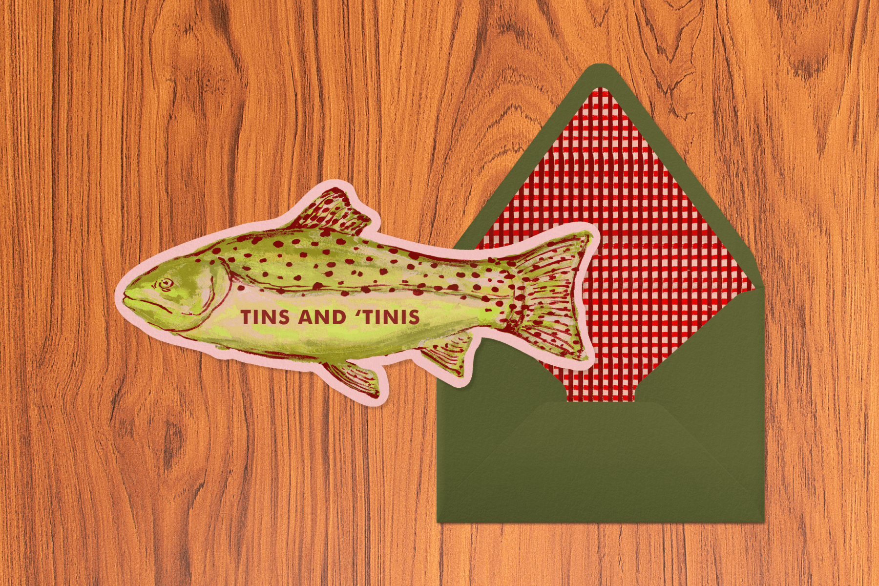 Green and red fish-shaped party invitation sits to the left of a dark green envelope with a red gingham interior against a light wood-textured backdrop.