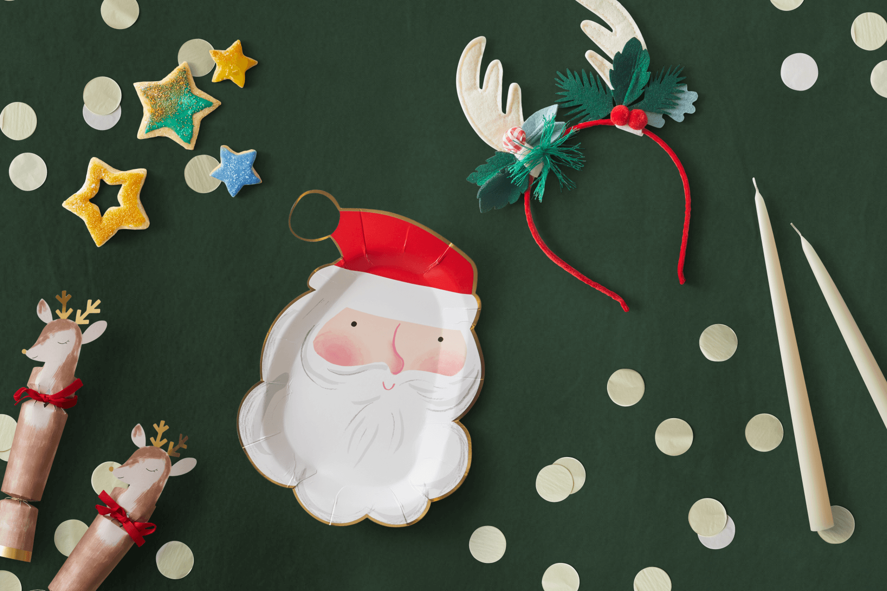 Overhead shot of Christmast party decor including reindeer crackers, sugar cookies, a Santa plate, a reindeer antlers headband, and white candles.