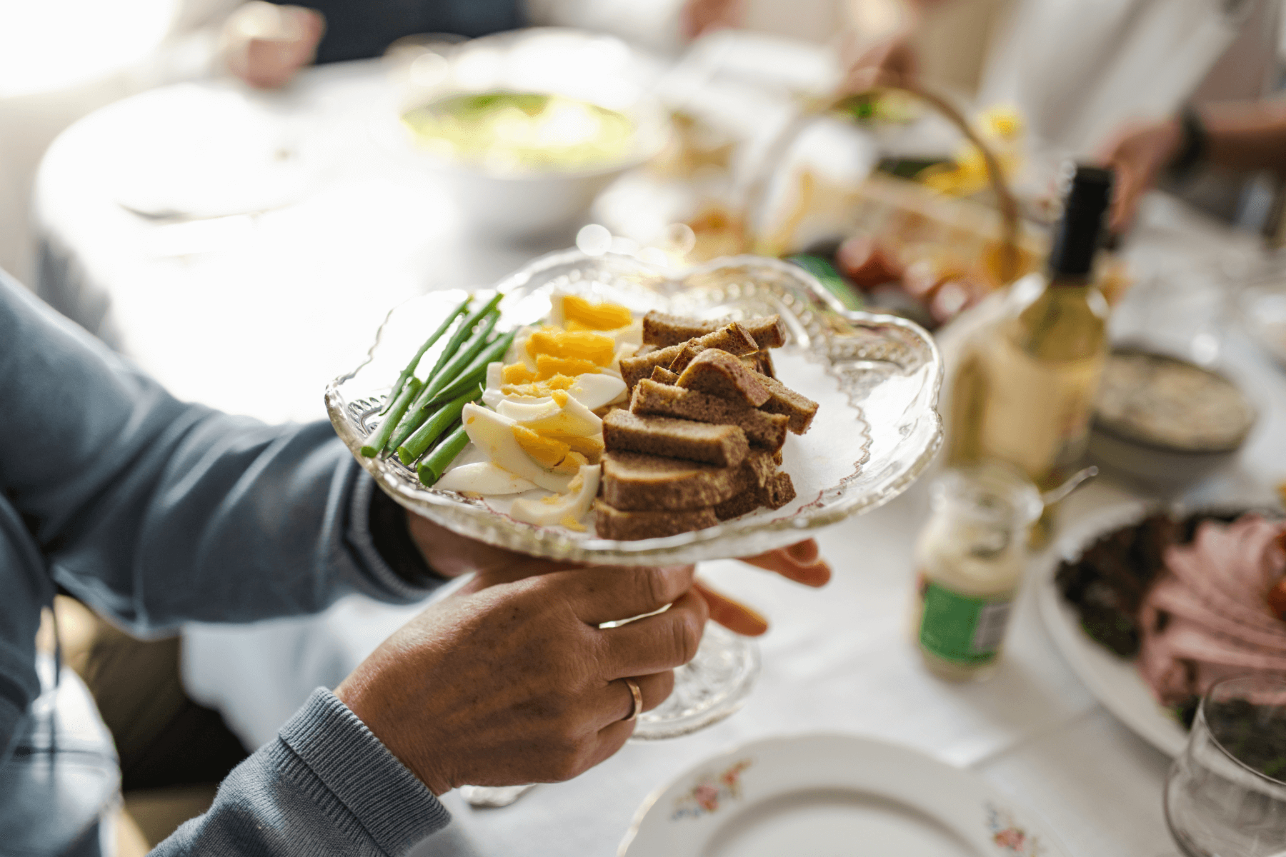Hands holding a glass tray of food including hard-boiled eggs, bread, and leeks. 
