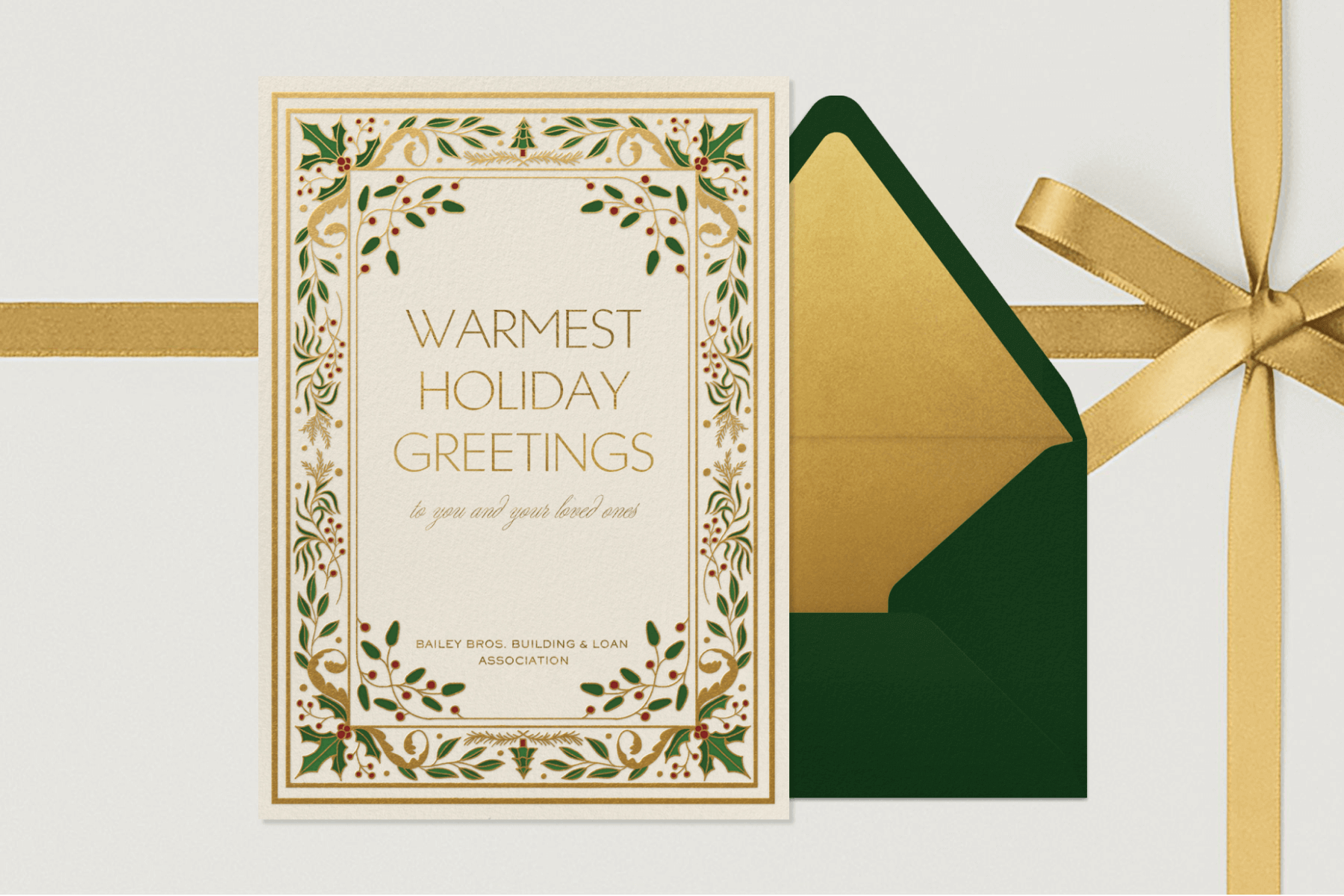 A holiday card with ornate gold and holly leaf border beside a green envelope with gold liner on a background with a gold ribbon bow.