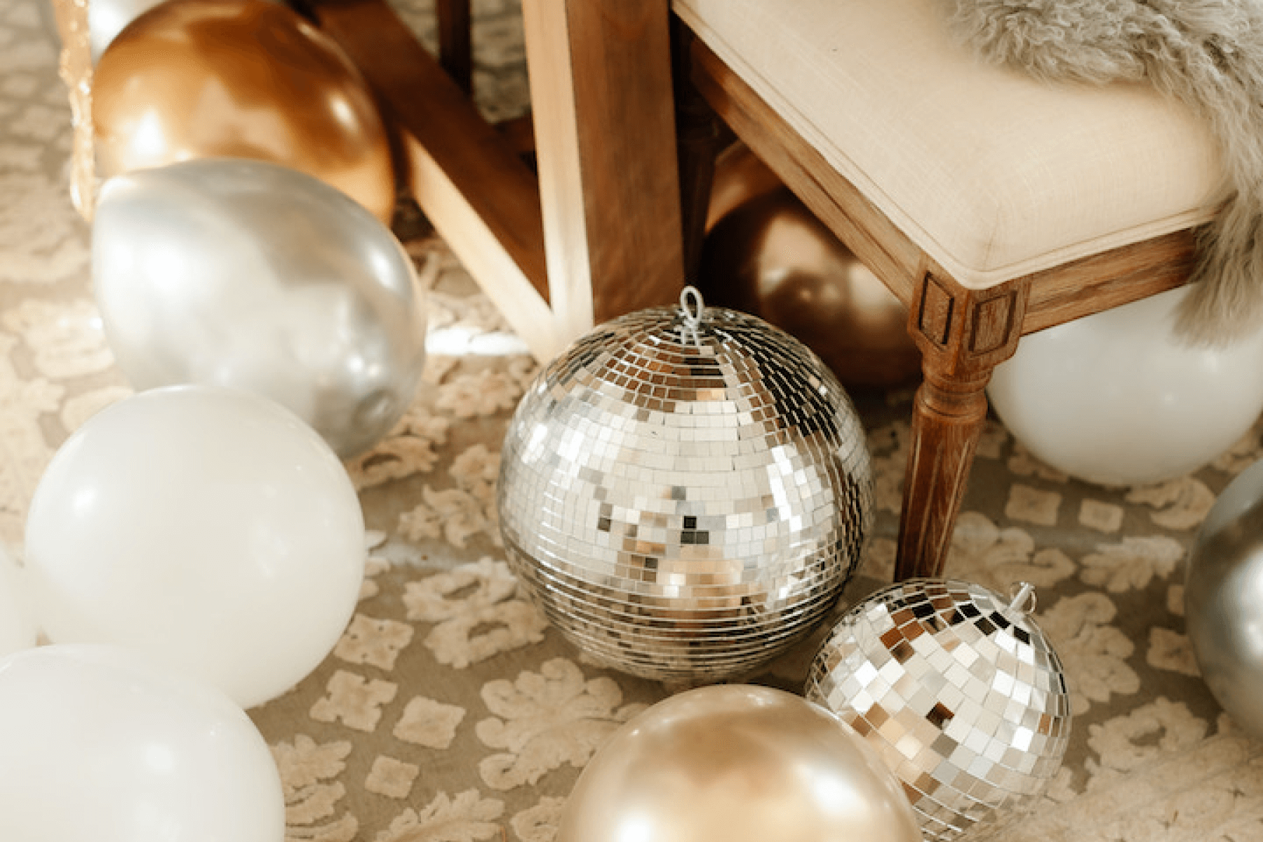 Two disco balls on the floor under a chair surrounded by white, silver, and gold balloons.