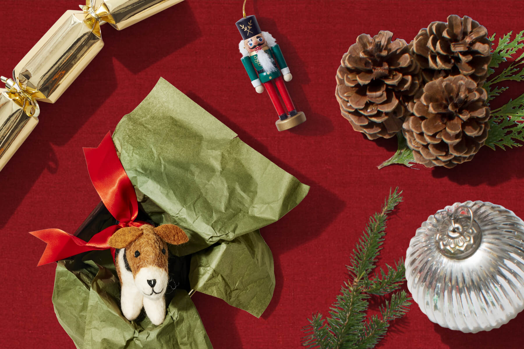 A collection of holiday gifts including a cracker, a felt dog ornament, a nutcracker ornament, and a bulb ornament, with greenery.